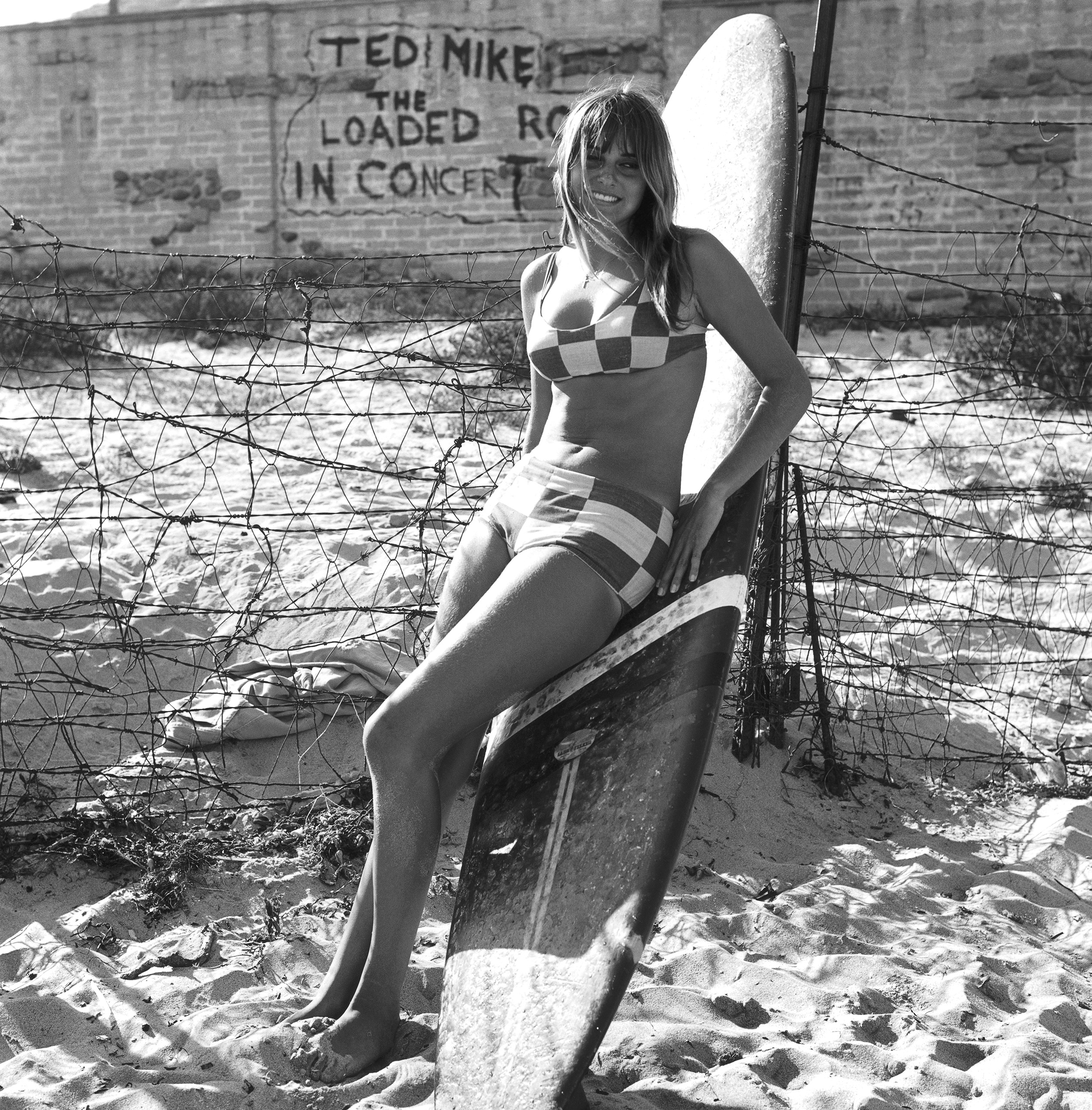 A woman in a checkered bikini leaning on a surf board Circa 1967 in Los Angeles.