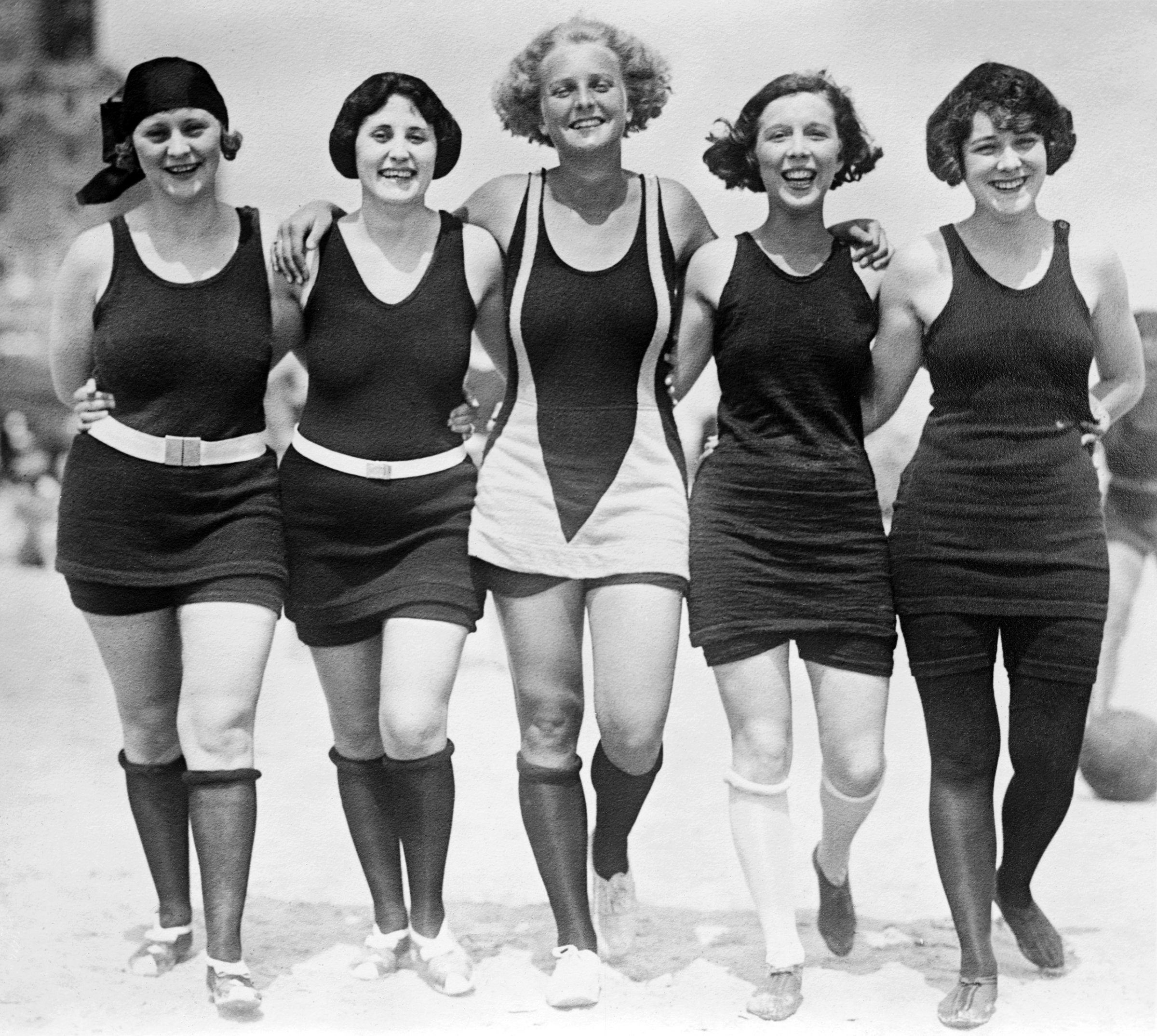 Five women walking arm-in-arm on the beach wearing wool bathing suits, circa 1925.