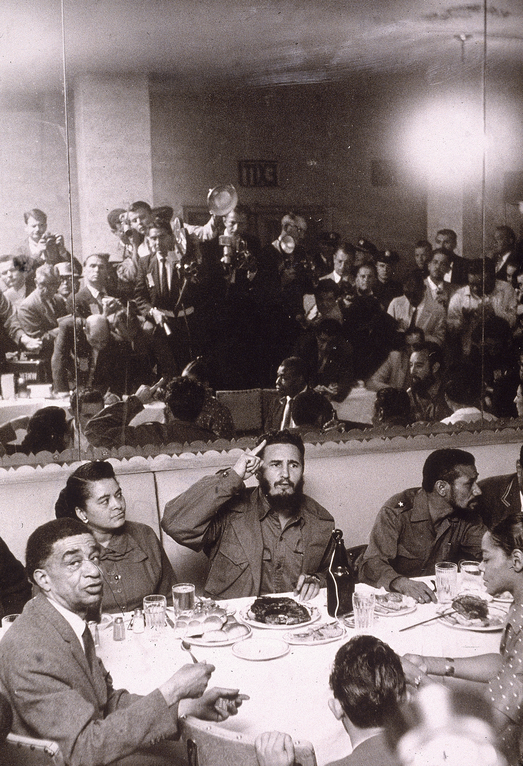 Cuban President Fidel Castro entertains a group of people at a dinner table in Harlem during a trip to New York City, as the press takes photographs in the background, 1959.