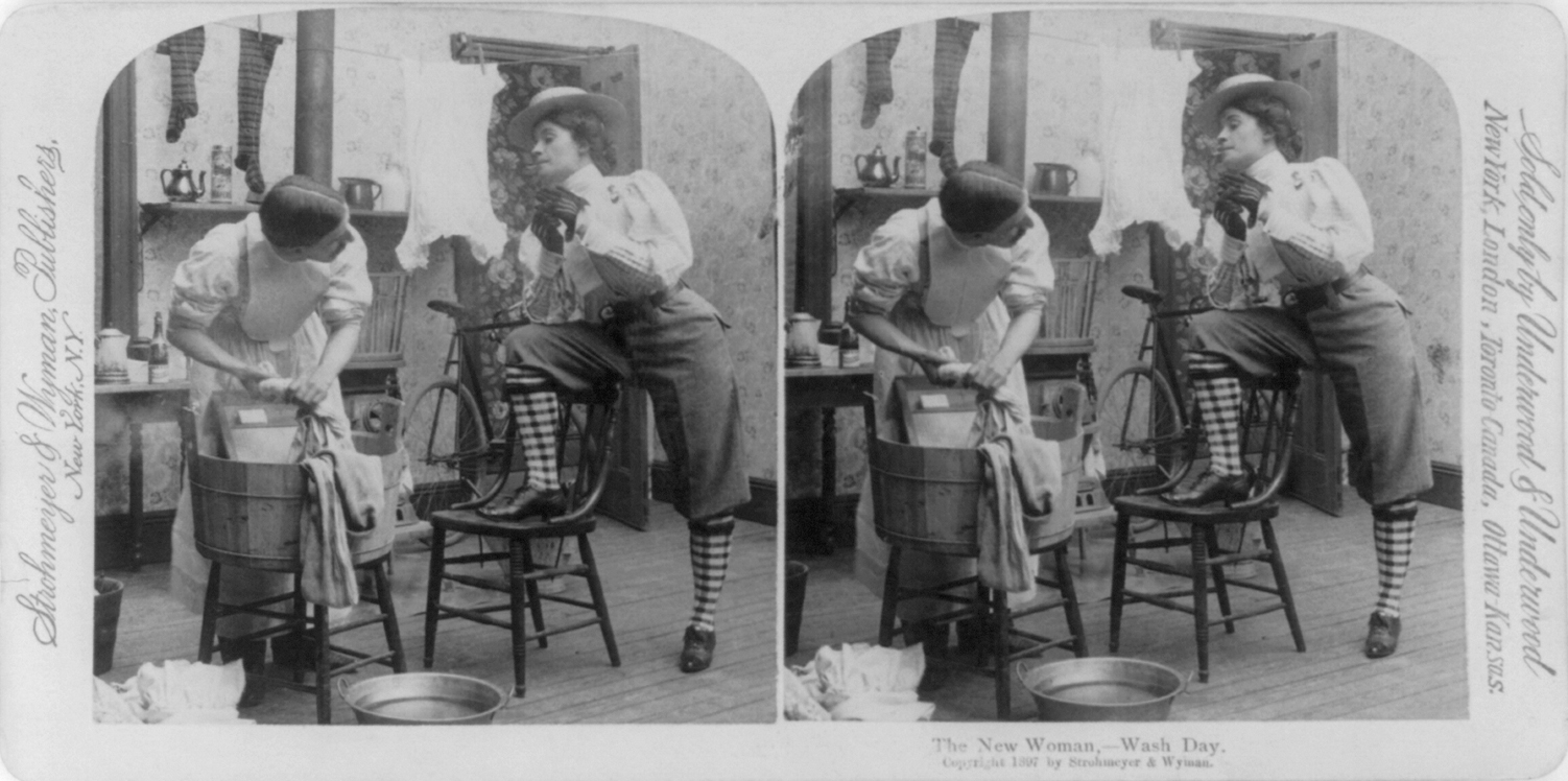 Stereograph card of "The New Woman" with cigarette in mouth, wearing pants, hat, and gloves, standing with one foot on chair and facing a man wearing a dress and doing laundry, circa 1897.