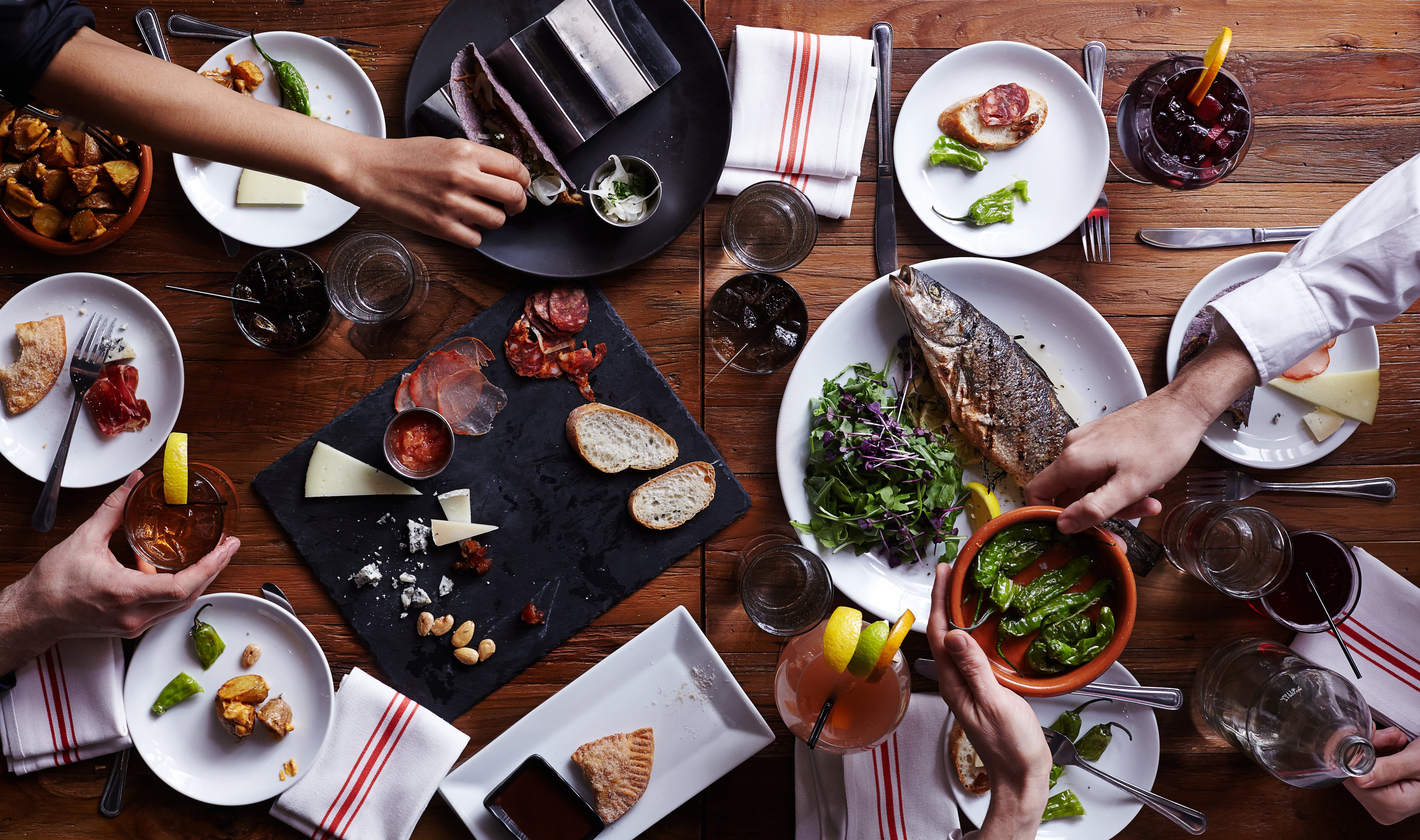 An aerial view of a group of people passing dinner plates with fish, charcuterie, and a variety of tapas and drinks. (Getty Images)