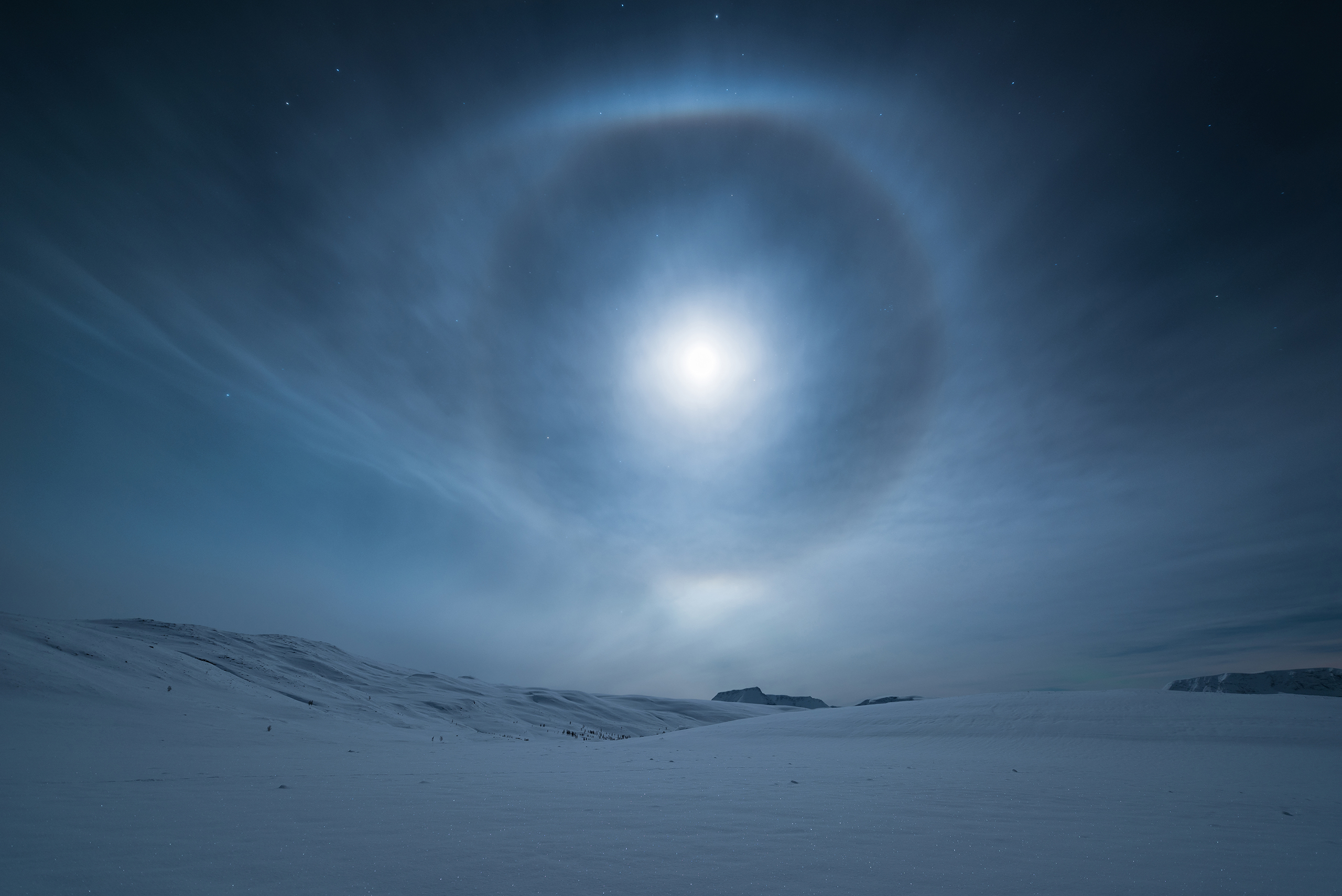 A mesmerizing lunar halo forms around our natural satellite, the Moon, in the night sky above Norway. The halo, also known as a moon ring or winter halo, is an optical phenomenon created when moonlight is refracted in numerous ice crystals suspended in the atmosphere.