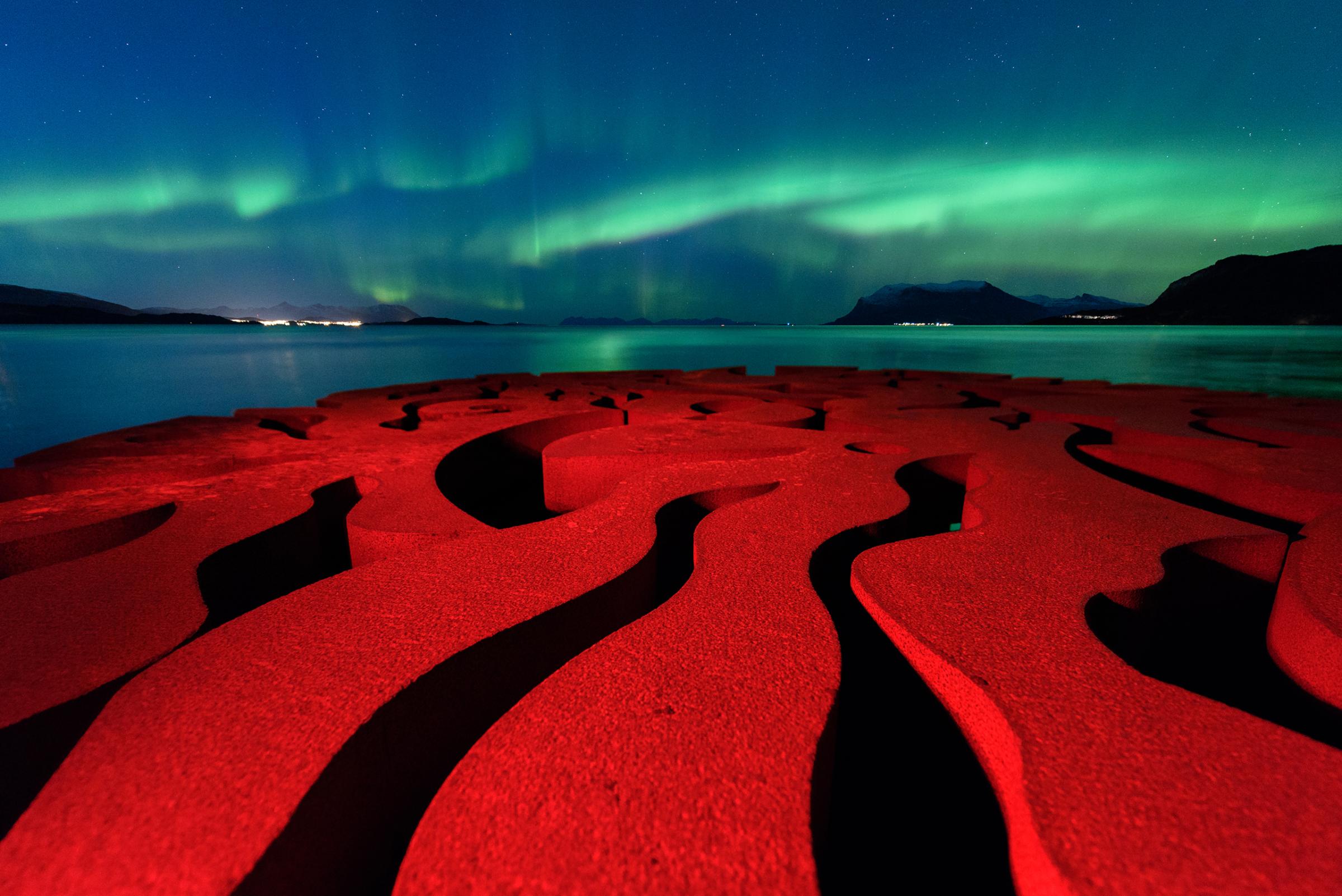 The rusty red swirls of the circular, iron sculpture, Seven Magic Points, in Brattebergan, Norway, mirror the rippling aurora above.