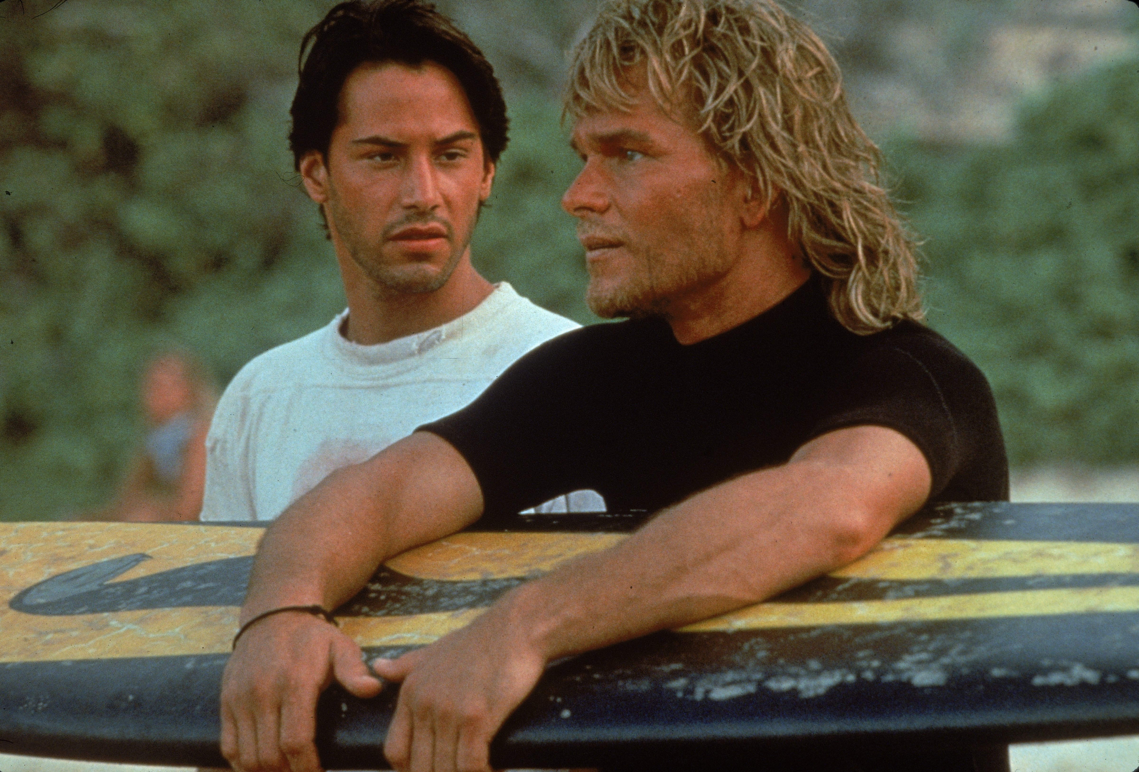Lebanese-born American actor Keanu Reeves and American actor Patrick Swayze stand on a beach as Swayze holds a surfboard during the filming of the action movie <i>Point Break</i> directed by Kathryn Bigelow, 1991. (Richard Foreman/Fotos International—Getty Images)