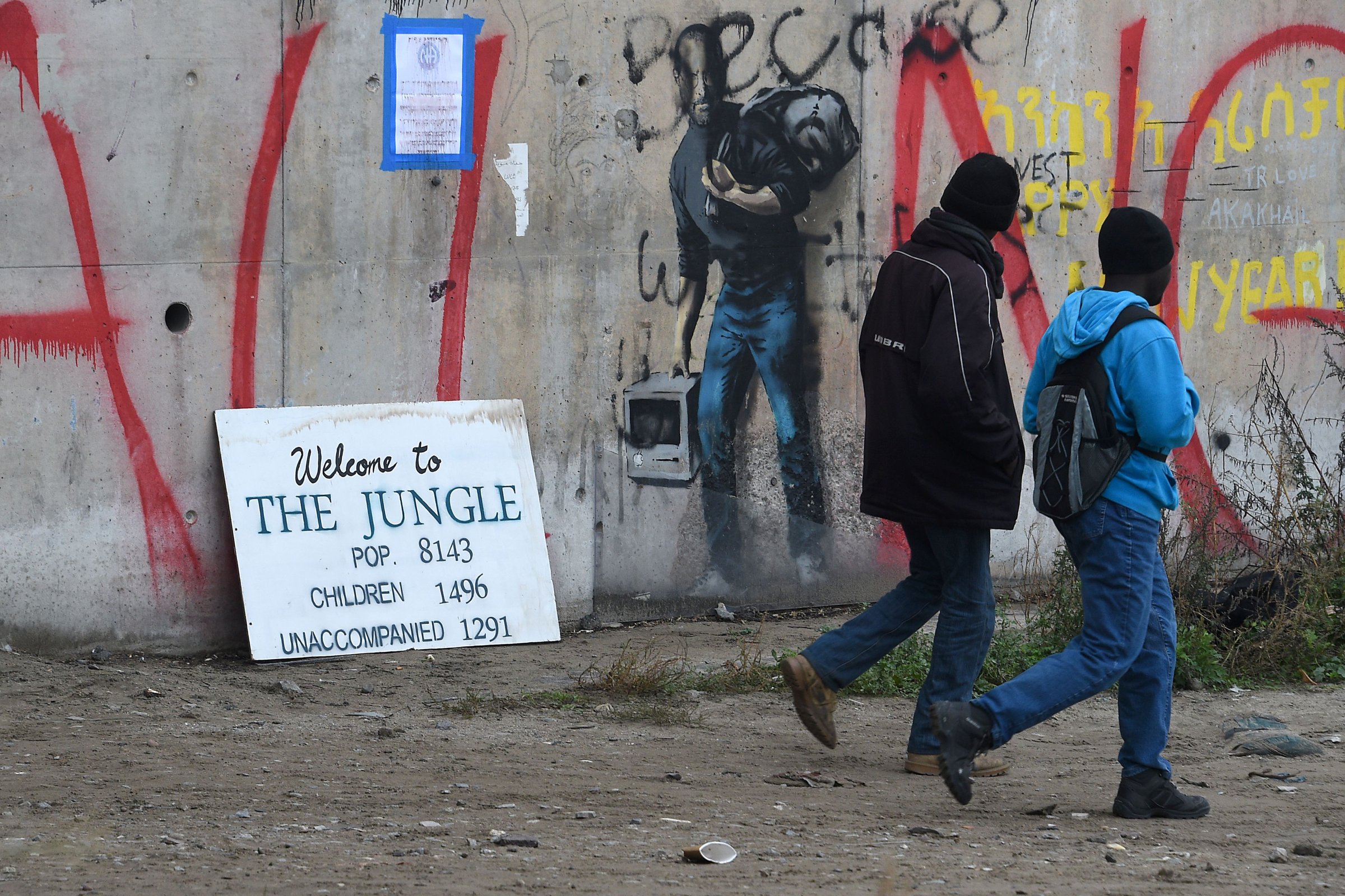 Migrants walk past a graffiti by street artist Banksy representing Apple founder Steve Jobs as a migrant during the full evacuation of the Calais "Jungle" camp, in Calais, northern France, on October 24, 2016.