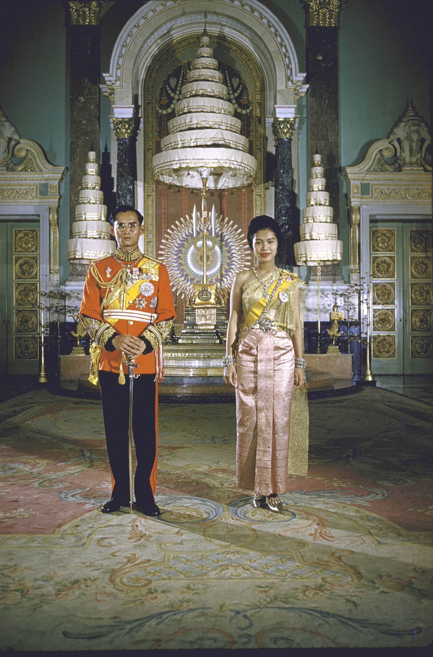 Formal portrait of King Bhumibol Adulyadej and Queen Sirikit at the Palace in 1960. The nine-tiered parasol in the background is a symbol of the Chakri Dynasty.