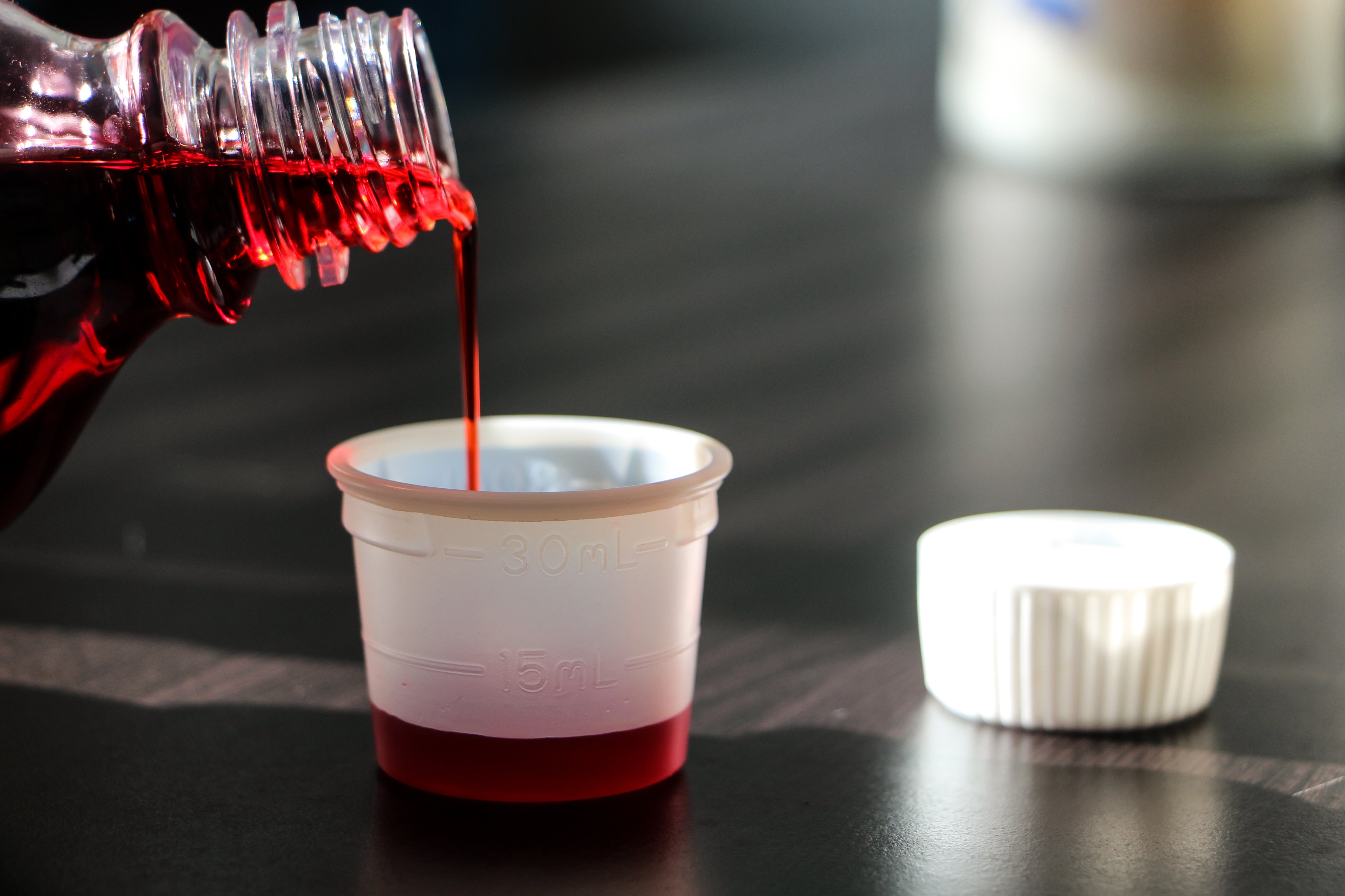 Close-Up Of Bottle Pouring Cough Syrup In Container On Table