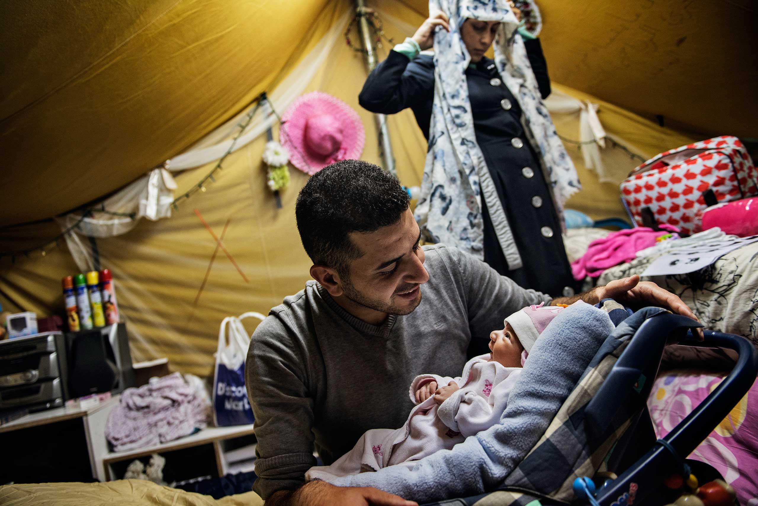 Yousef Alarsan holds his newborn baby daughter Rahaf, while his wife Nourelhuda, stands behind them in their tent in the  Oreokastro camp, Nov. 8, 2016.   In 2014, Yousef left school in Deir ez-Zor and moved to a small nearby town to escape induction in the Syrian Army.  Once ISIS took control of the town, fearing for his life, he and Nour married and left Syria. Rahaf was born on Nov. 1, 2016.