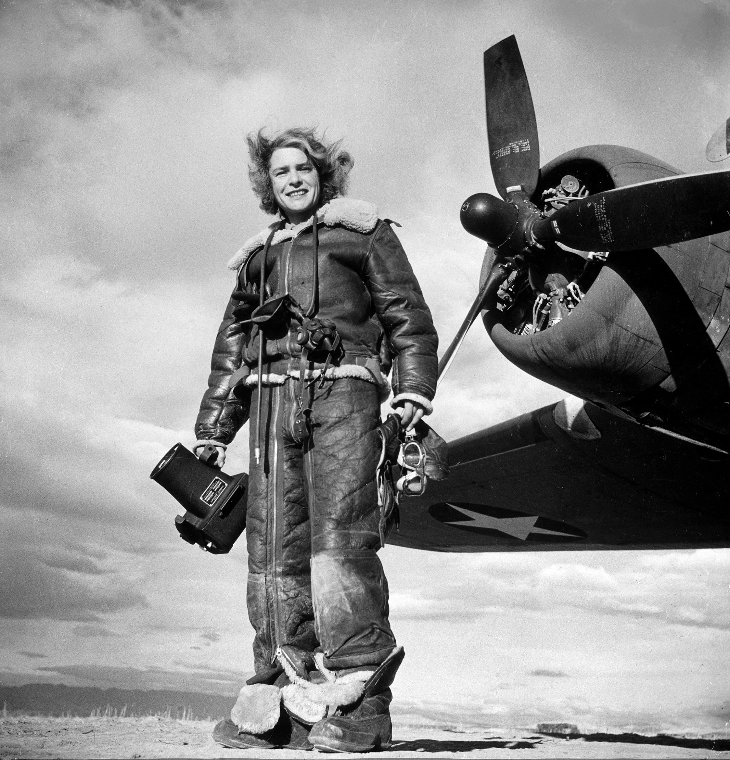 LIFE photographer Margaret Bourke-White clad in fleece flight suit while holding aerial camera, standing in front of Flying Fortress bomber in which she made combat mission photographs of the US attack on Tunis, 1943.