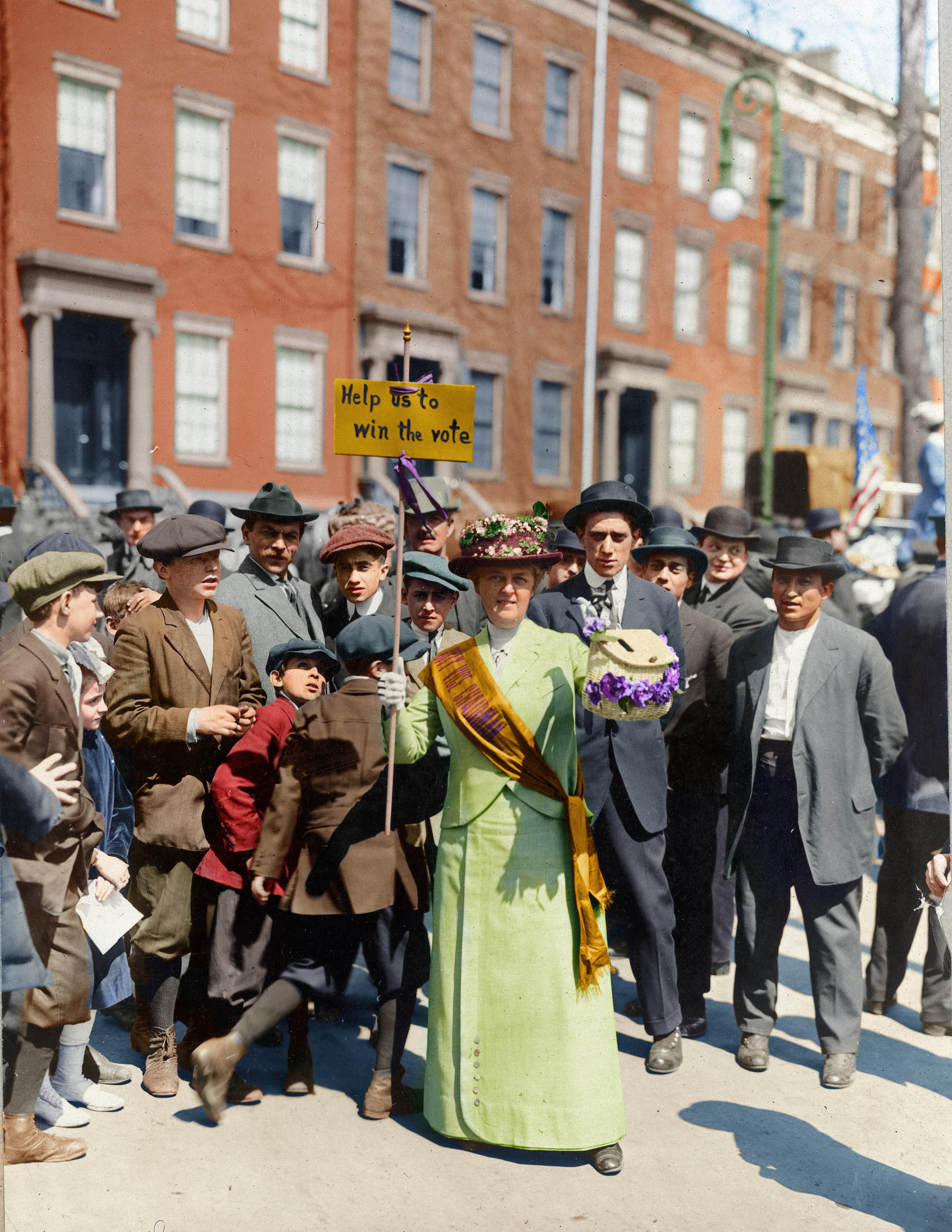 Mrs. Suffern wearing a sash and carrying a sign that says "Help us to win the vote," surrounded by a crowd of men and boys at a Women's Suffrage Parade in 1914.