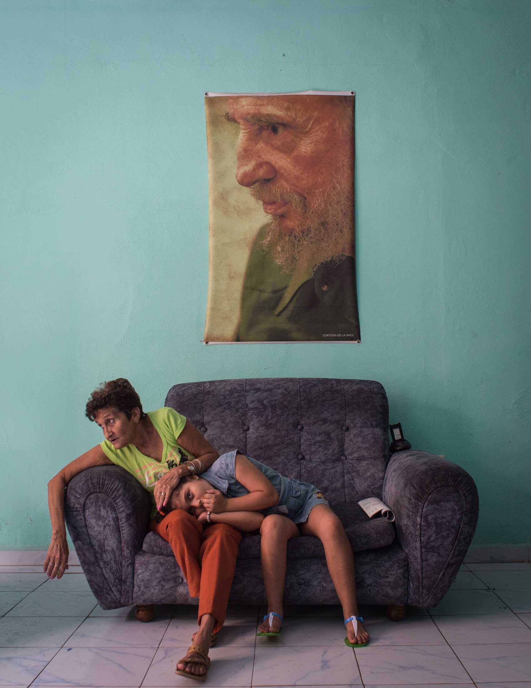 A woman strokes a girl’s head as she rests on her lap whilst sitting on a sofa in a police station in Camaguey, Cuba, on Feb. 12, 2016, with a portrait of Fidel Castro hanging above them.