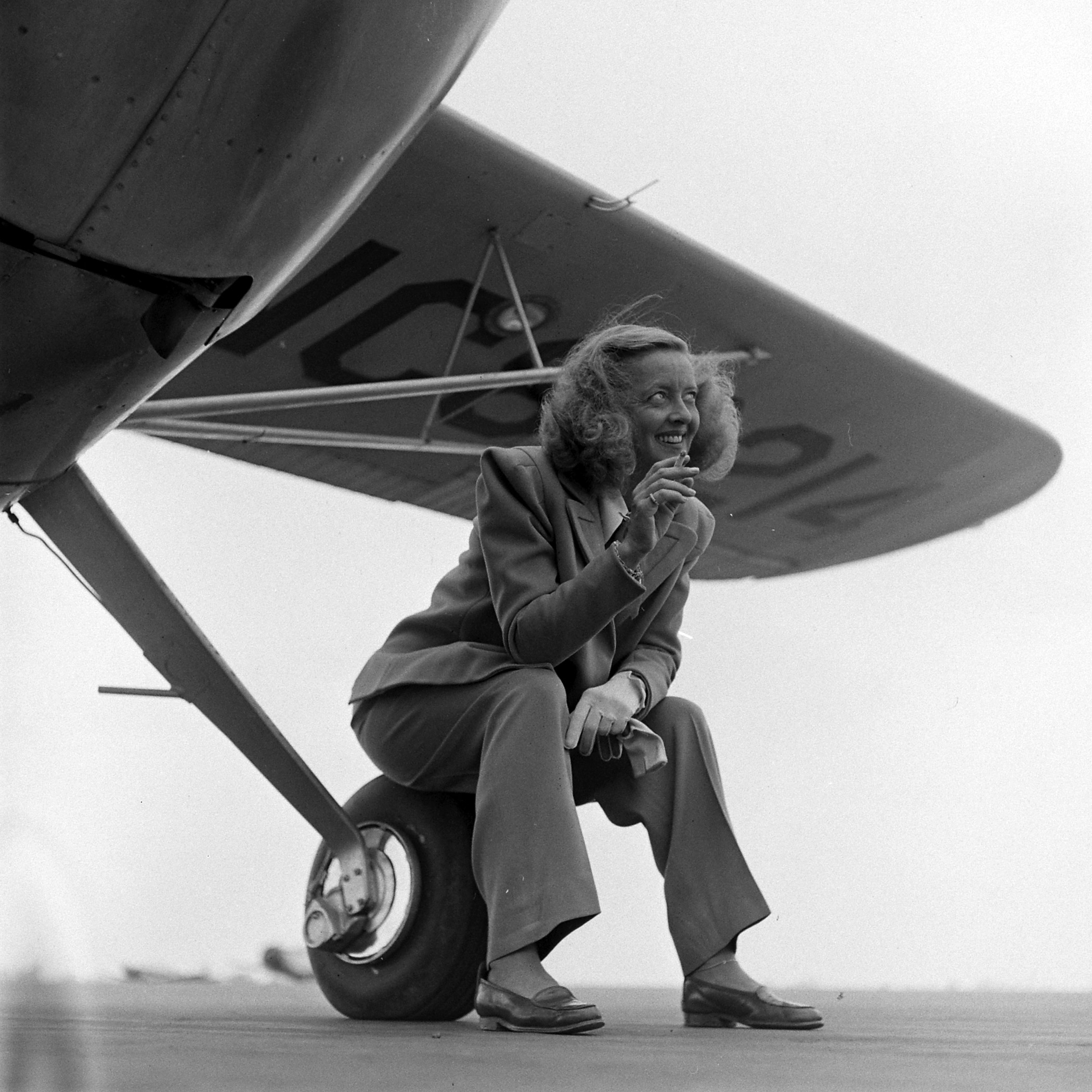 Bette Davis smoking and sitting on the wheel of a plane, 1947.