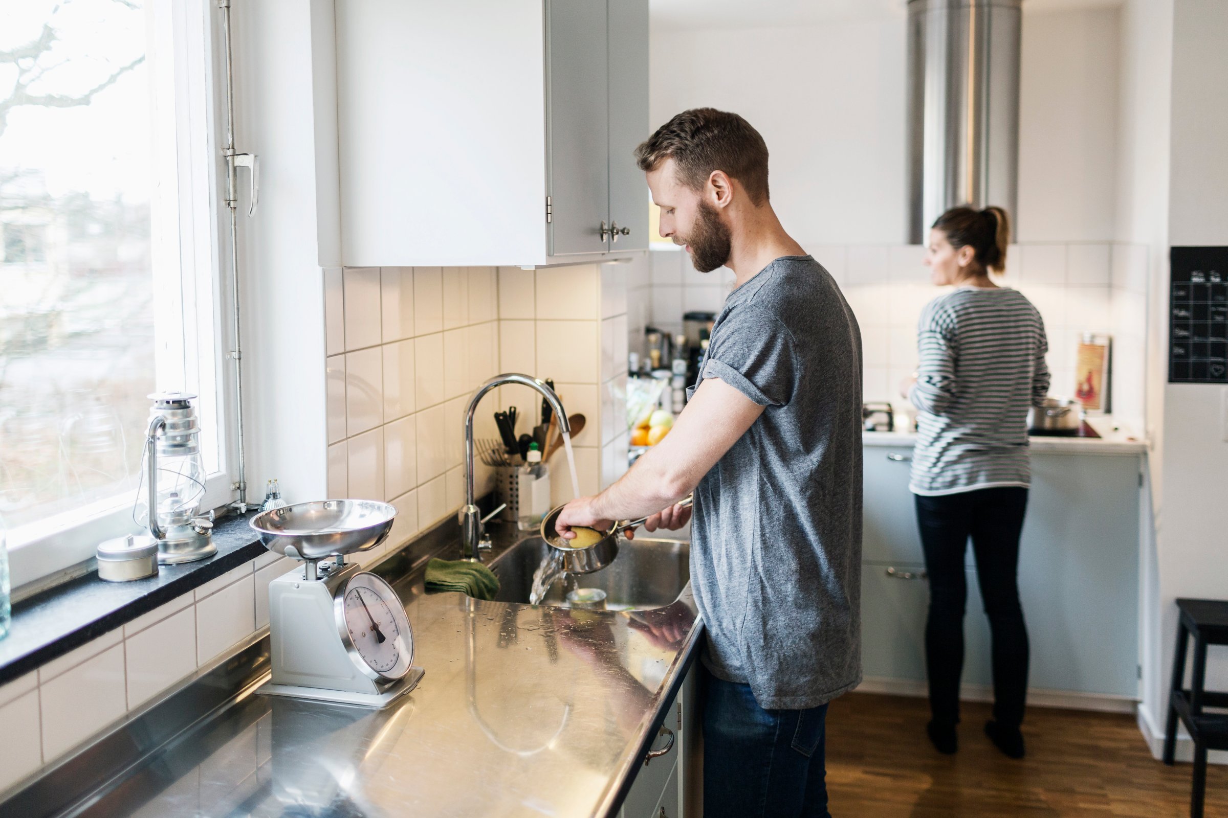 Man washing sauce pan while woman standing in background in kitchen
