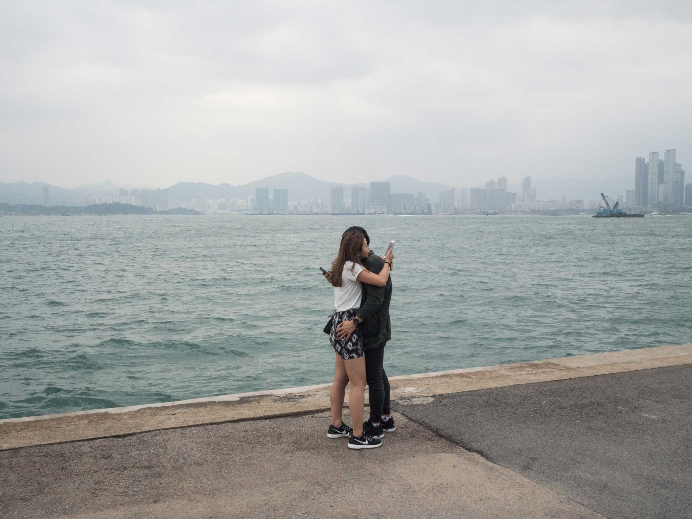 Hong Kong (Hong Kong SAR, China); Date (10, 30, 2016); two young lovers embrace in a hugh while checking their phones. "Instagram pier" is a public cargo pier located on the west side of Hong Kong island; here locals gather for fishing, excercising or walking their pets. More recently the Pier has raised to fame as the “instagram Pier”, whereyoung Hong Kongers and instagrammers come for selfies and scenic photos. I have recently begun curating its "instagram" dedicated account (@insta_pier).