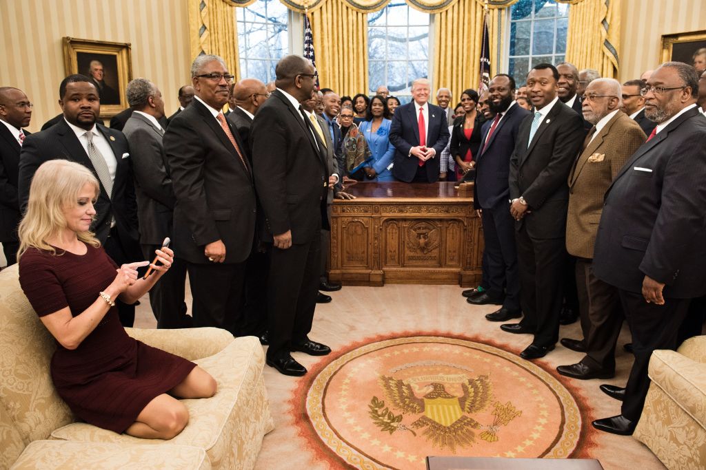 Kellyanne Conway (L) checks her phone after taking a photo as President Donald Trump and leaders of historically black universities and colleges pose for a group photo in the Oval Office of the White House on Feb. 27, 2017 in Washington, DC.