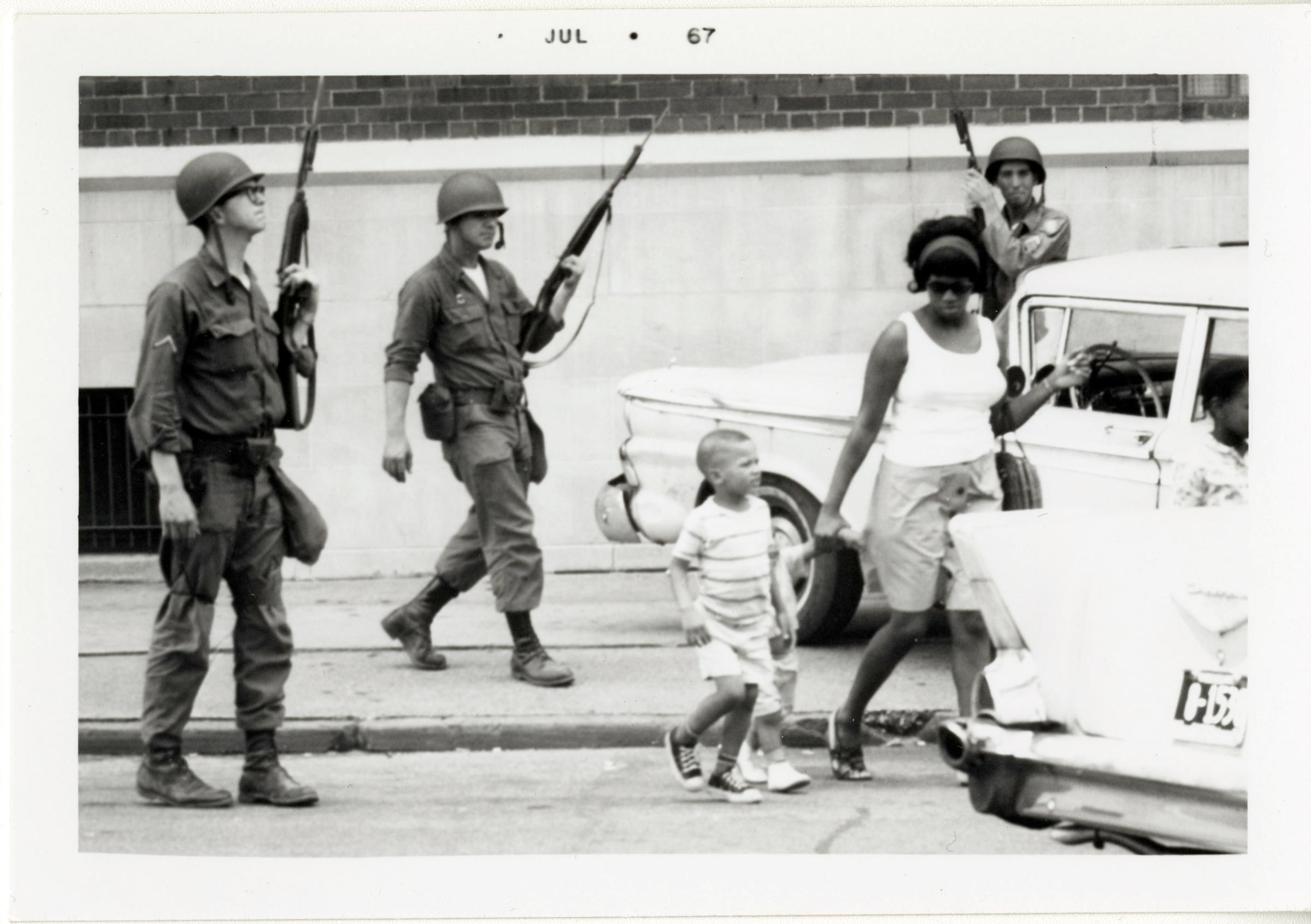 A woman and her children carry on with their daily activities as Michigan National Guardsmen patrol the streets in Detroit.