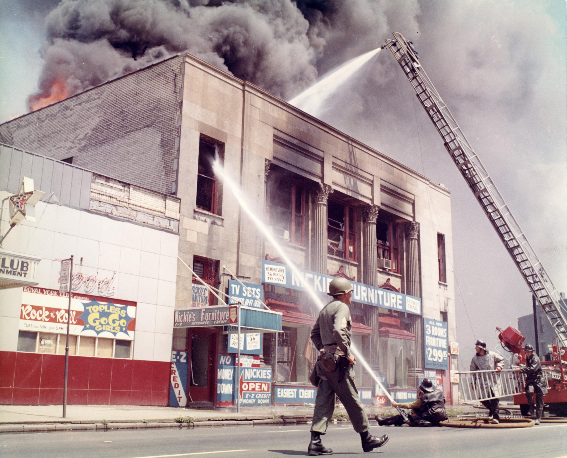 A U.S. Army soldier patrols the streets as Detroit firefighters suppress flames that have broken through the rooftop of a neighborhood furniture store.