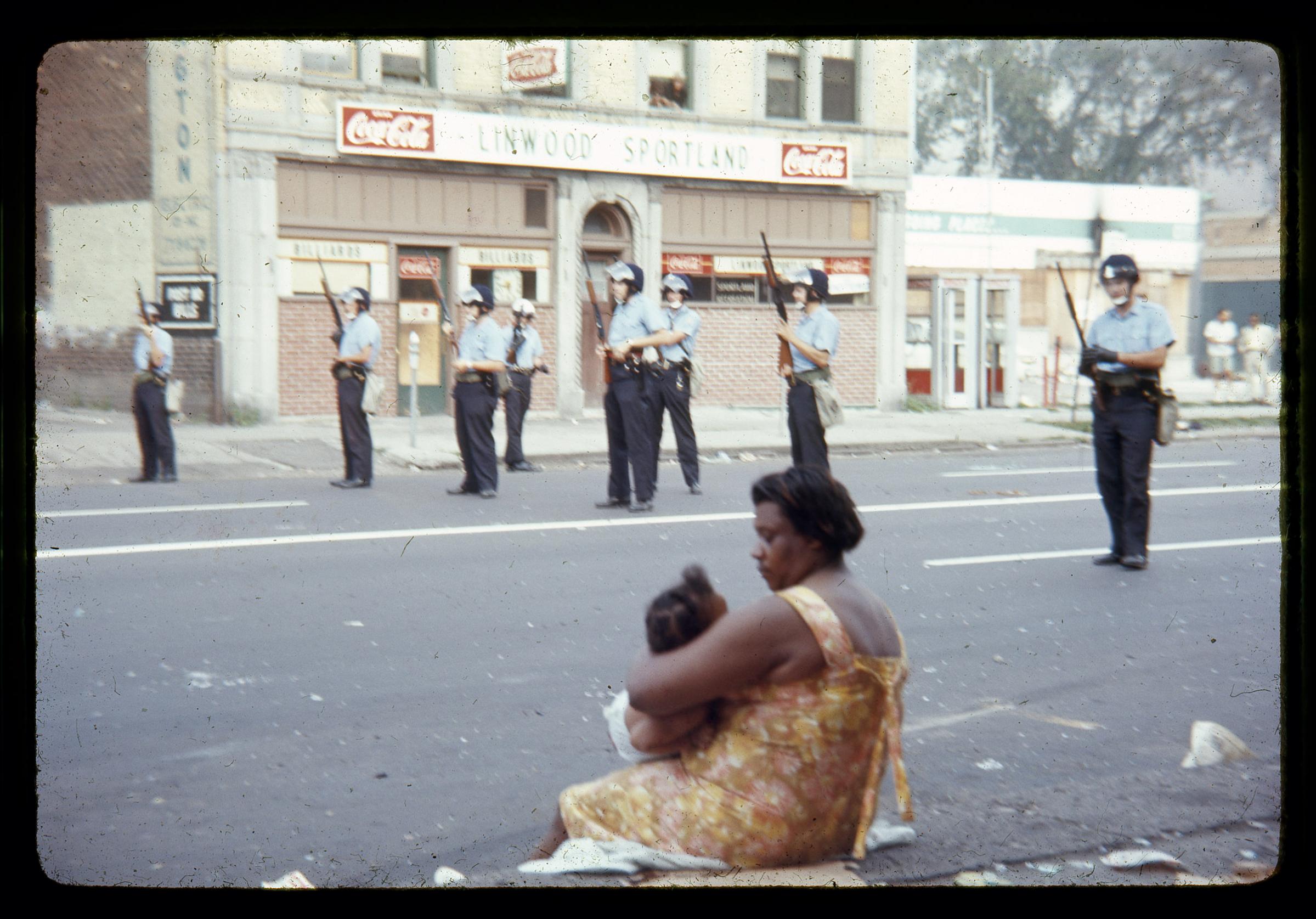 Several Detroit police officers clad in riot gear guard the streets of Detroit as a woman and child look on.