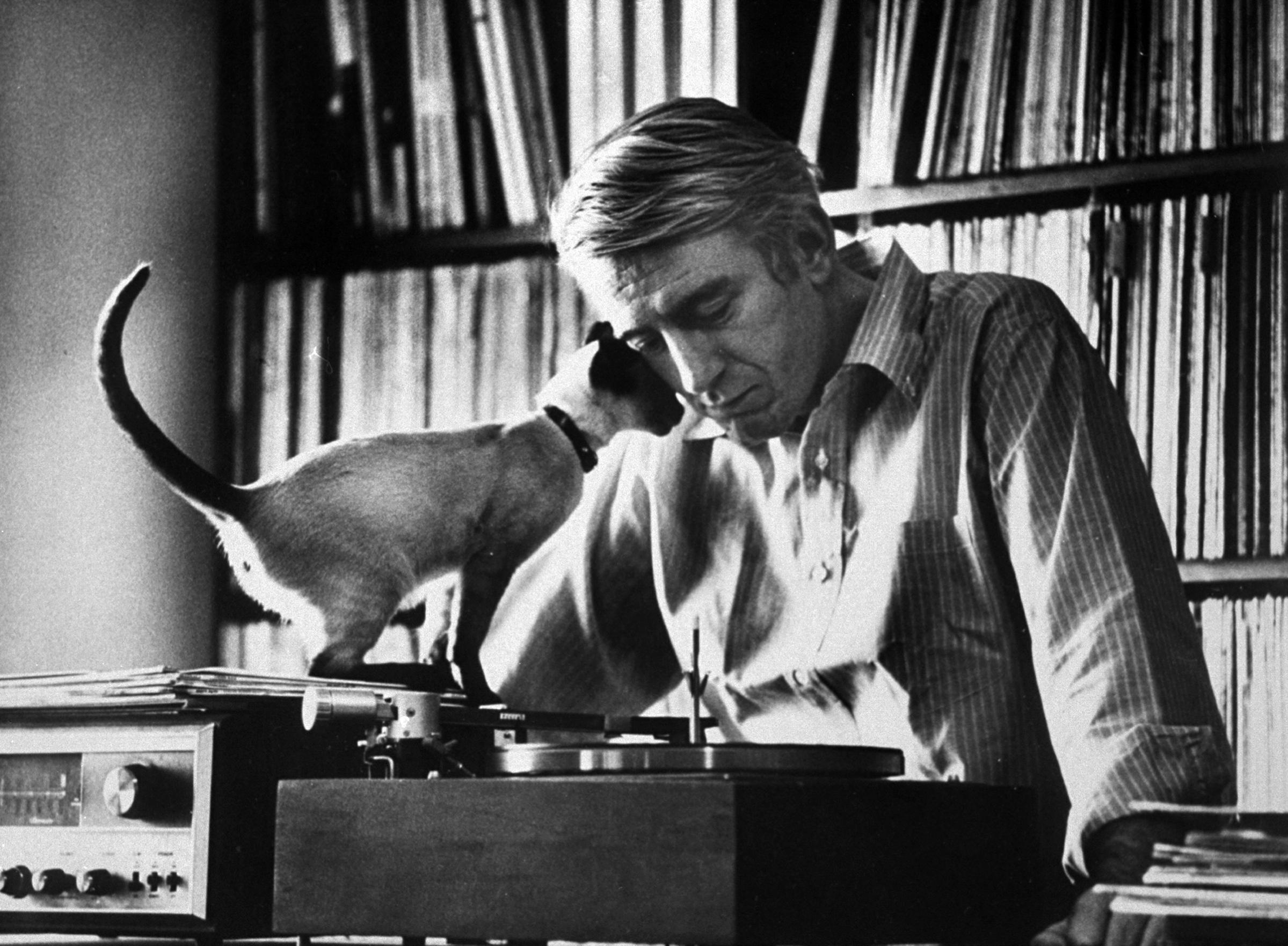 Poet Rod McKuen playing record on stereo set while pet Siamese cat nuzzles his face affectionately, 1967.