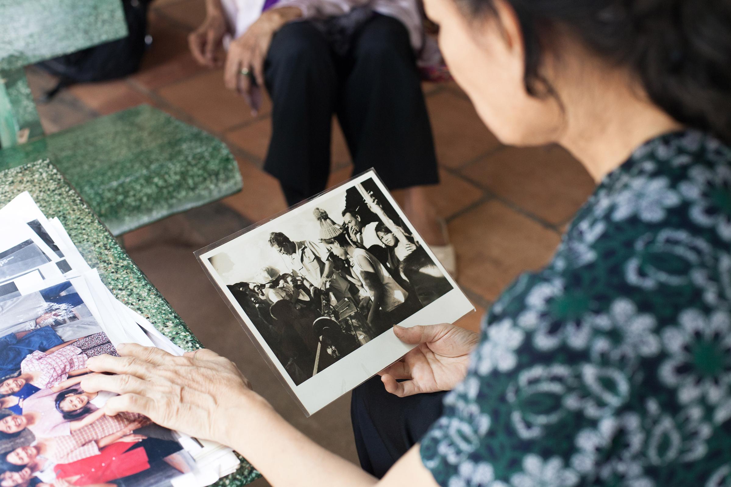 Nguyễn Thị Tròn (Nguyen Thi Tron) holds a laminated photograph from 1969 depicting her (in woollen hat), Vietnam War photographer Larry Burrows (wearing glasses), her father (at right) and her village neighbours when Burrowed delivered to her the gift of a sewing machine. This new picture was taken outside Nguyễn's small convenience store in Suoi Da commune in Duong Minh Chau district, Tây Ninh province, Vietnam.