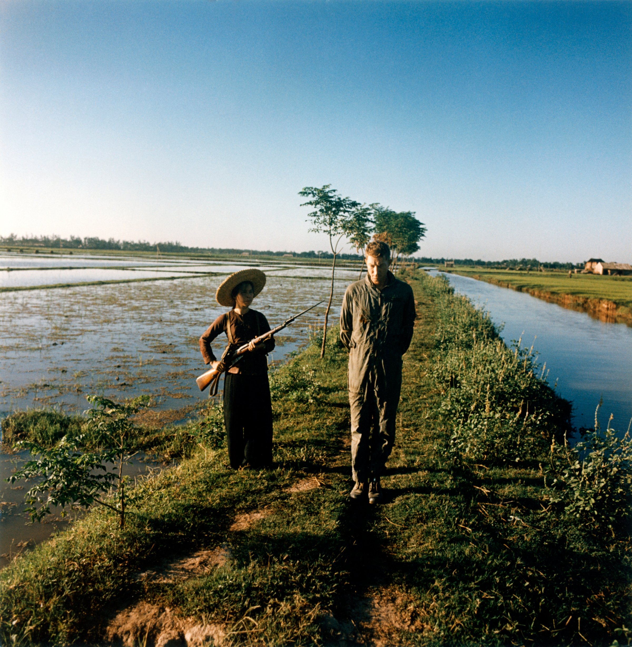 The captured US pilot major Pewey Waddell is being guarded by a militiawoman with a gun and a bayonet on a rice field - 1967
