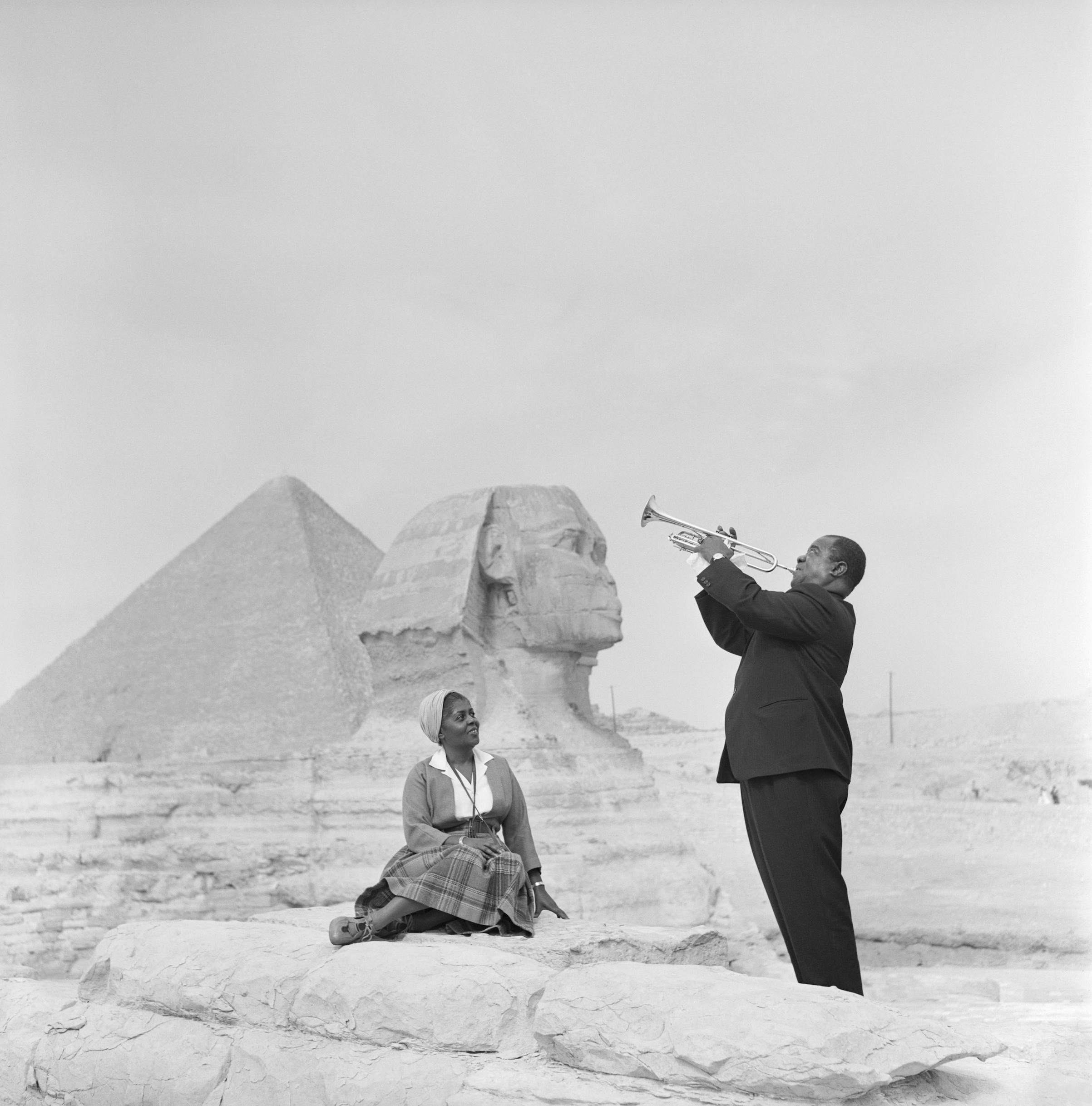 American jazz trumpeter Louis Armstrong plays the trumpet while his wife sits listening, with the Sphinx and one of the pyramids behind her, during a visit to the pyramids at Giza. (Bettmann / Getty Images)