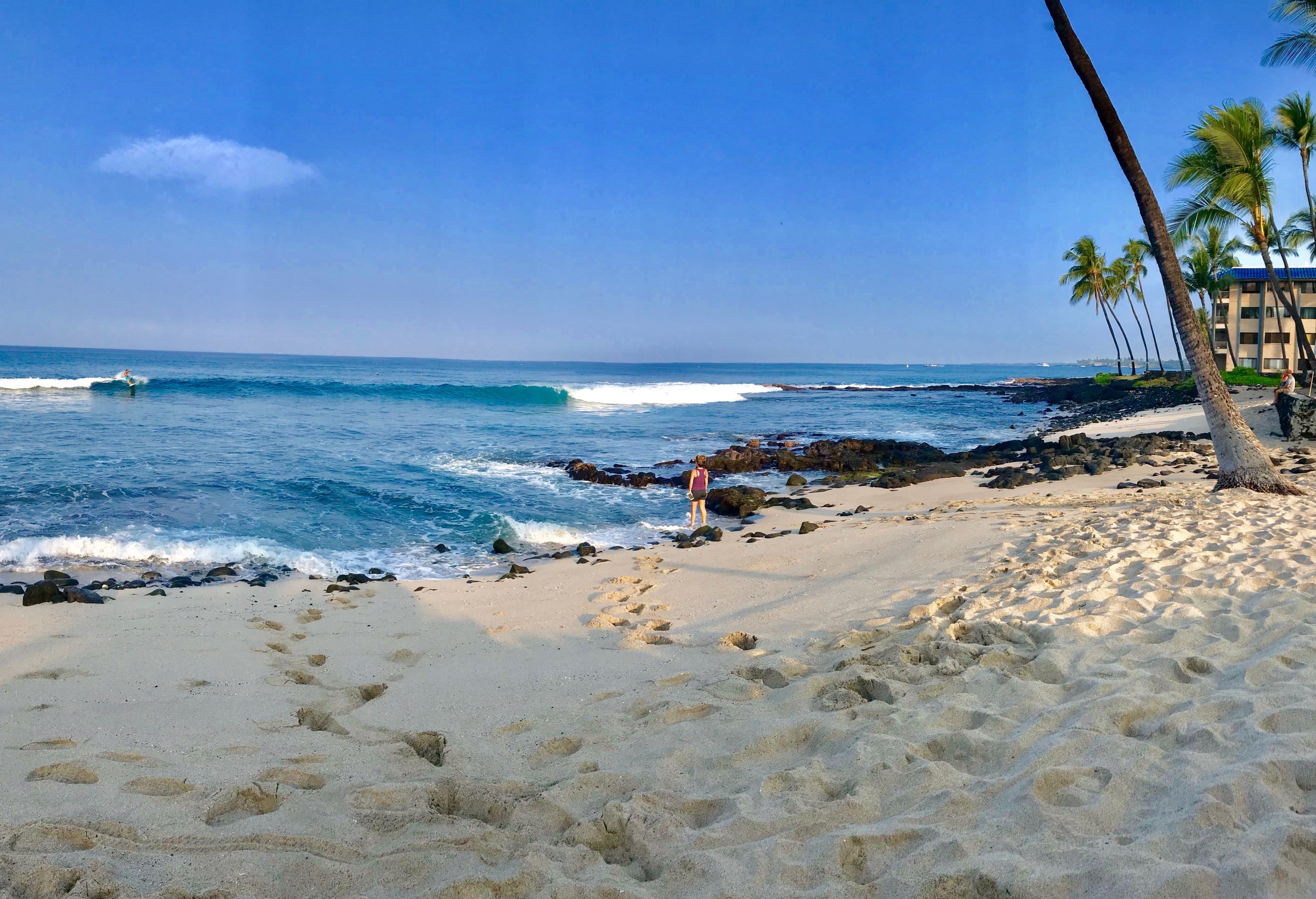 The Kona shore is mostly empty with only one surfer in the bright blue ocean.