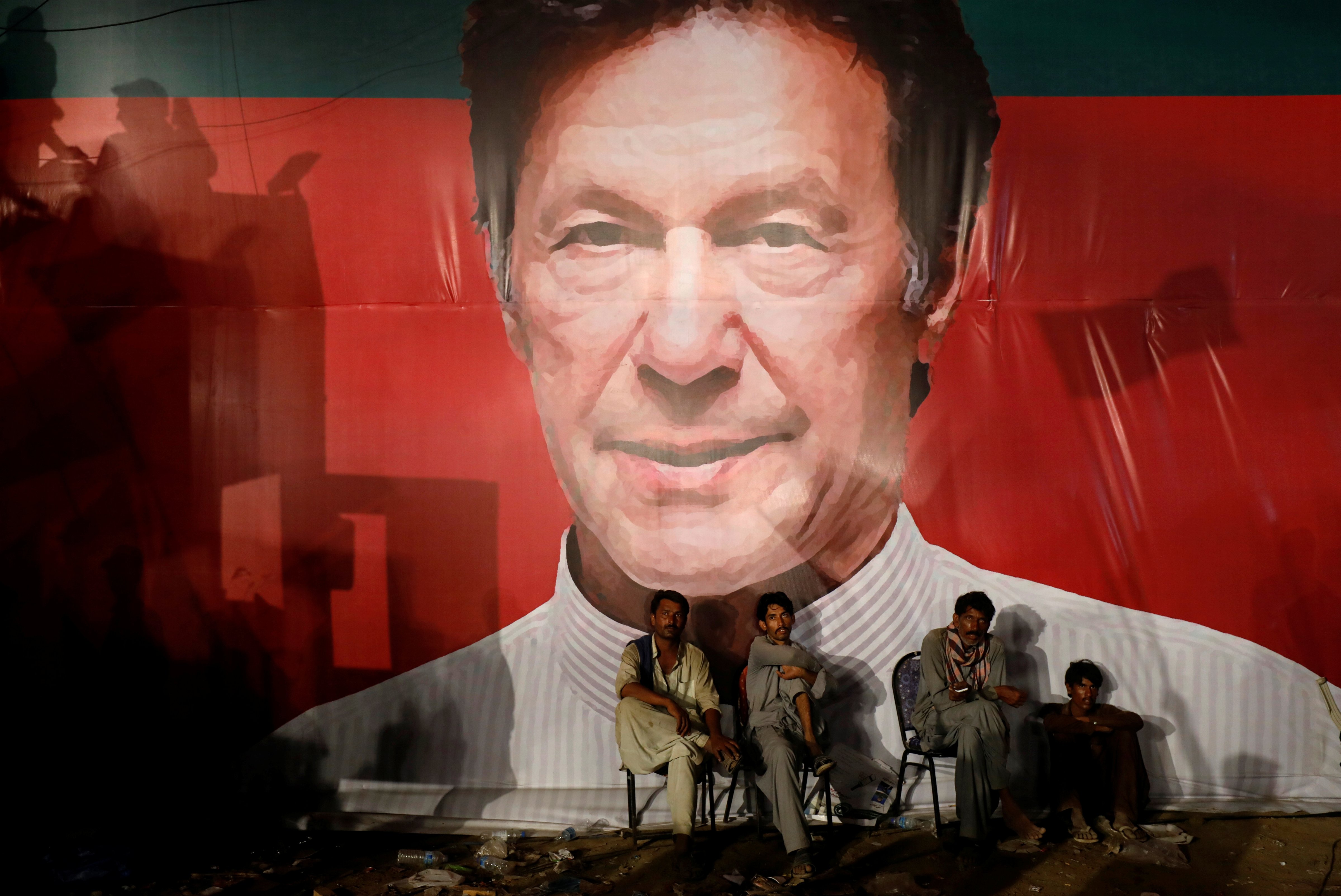 Labourers, who set up the venue, sit under a wall with a billboard displaying photo of Imran Khan, chairman of the Pakistan Tehreek-e-Insaf (PTI), political party, as they listen to him during a campaign rally ahead of general elections in Karachi
