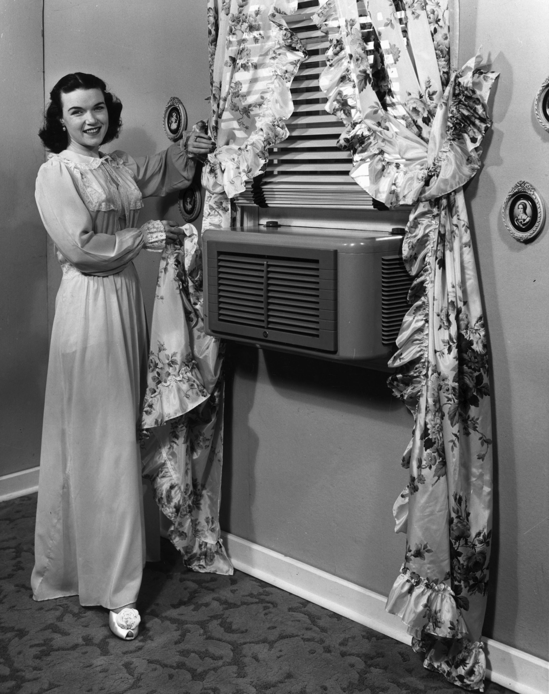 circa 1945:  A woman dressed in a long nightgown pulls a ruffled floral print curtain back to show a modern air conditioner mounted in the window. (Lambert/Getty Images)