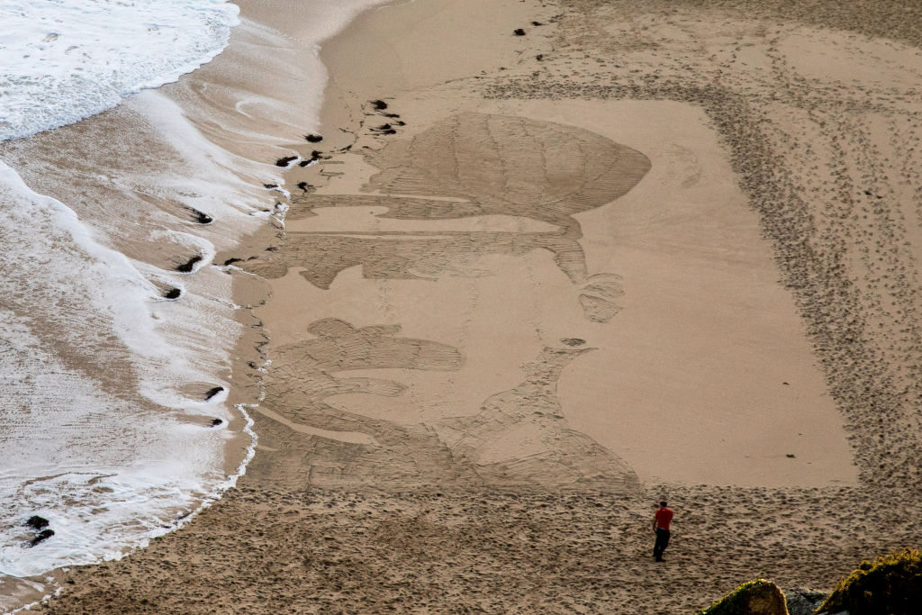 People on Porthcurno beach watch as waves wash away a large scale sand portrait of Lieutenant Richard Charles Graves-Sawle, one of a number from the project called Pages of the Sea near Penzance on November 11, 2018 in Cornwall, United Kingdom. (Matt Cardy—Getty Images)