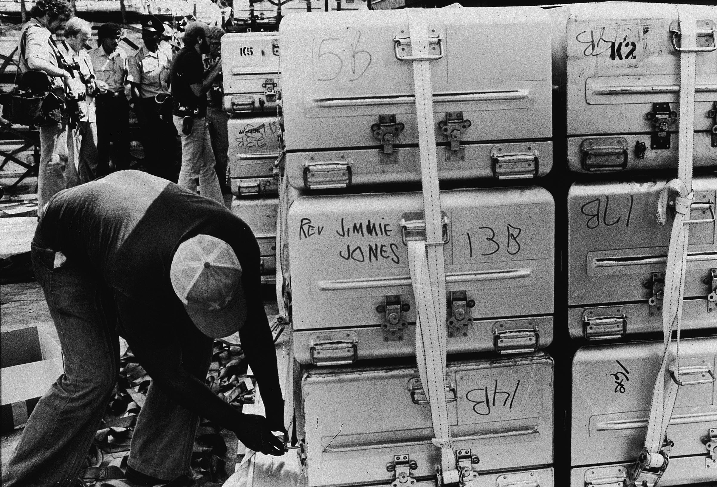 An unidentified man a strap onto a stack of aluminum coffins for shipment to the United States, following the more than 900 deaths in the mass suicide staged in Jonestown by members of the People's Temple and their leader, the Reverend Jim Jones, Georgetown, Guyana, Nov. 23, 1978. A group of photographers and police officers stand in the background. (New York Times Co.—Getty Images)