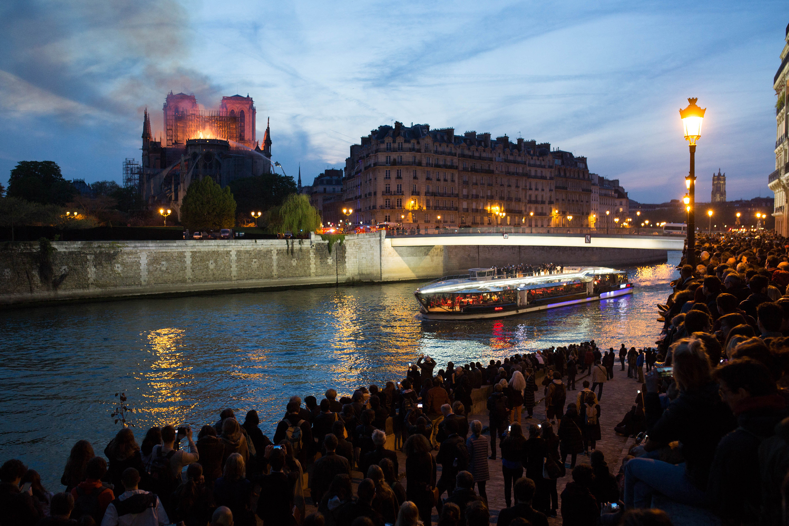 Bystanders look on as flames and smoke are seen billowing from the roof at Notre-Dame Cathedral with the Seine and boats by sun set river in Paris on April 15, 2019. (Raphael Lafargue—Abaca/Sipa USA)