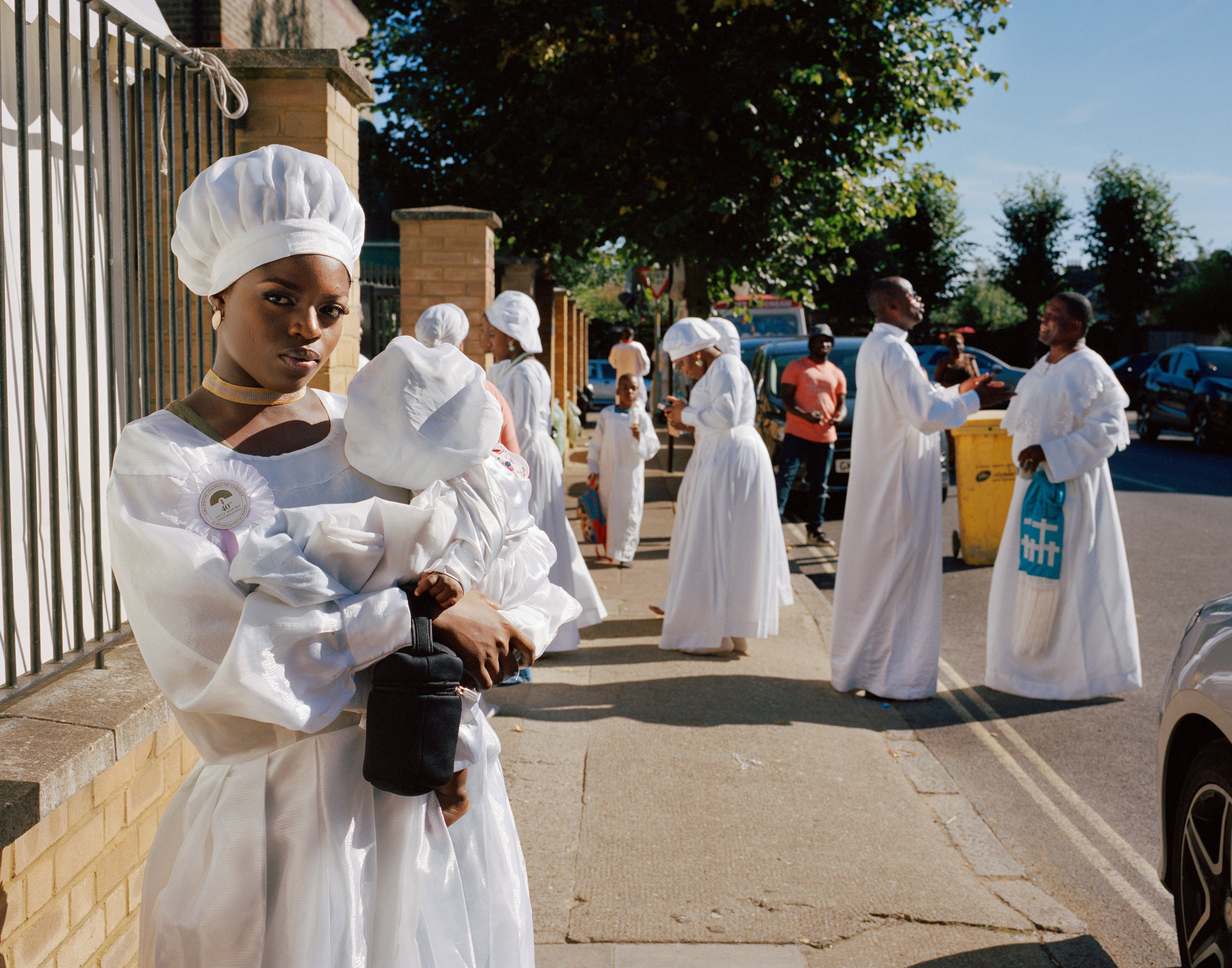 Hannah stands on the street holding her baby cousin whilst members of the Congregation greet each other and chat in the background of the street. (Sophie Green)
