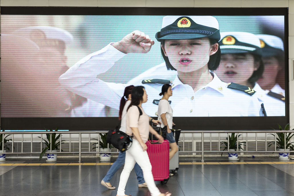 Travelers walk past an advertisement for the People's Liberation Army (PLA) on a screen near the Luohu border crossing in Shenzhen, China, on Aug. 4, 2019. (Qilai Shen&mdash;Bloomberg/Getty Images)