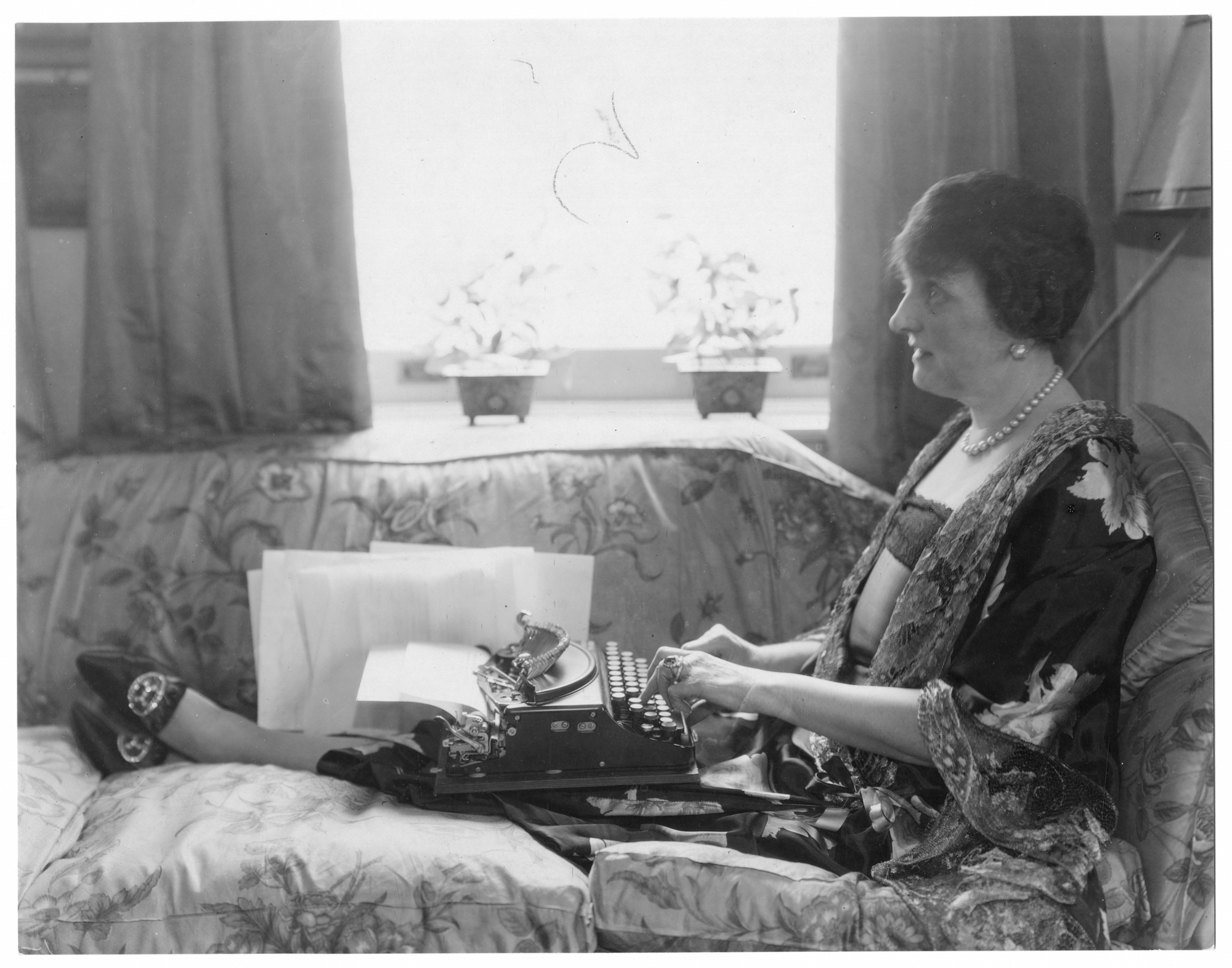 Etiquette author Emily Post, in pearls and flowing robe, sitting lengthwise on couch with legs stretched out, typewriter in her lap, 1927. (Jessie Tarbox Beals/The New York Historical Society/Getty Images)