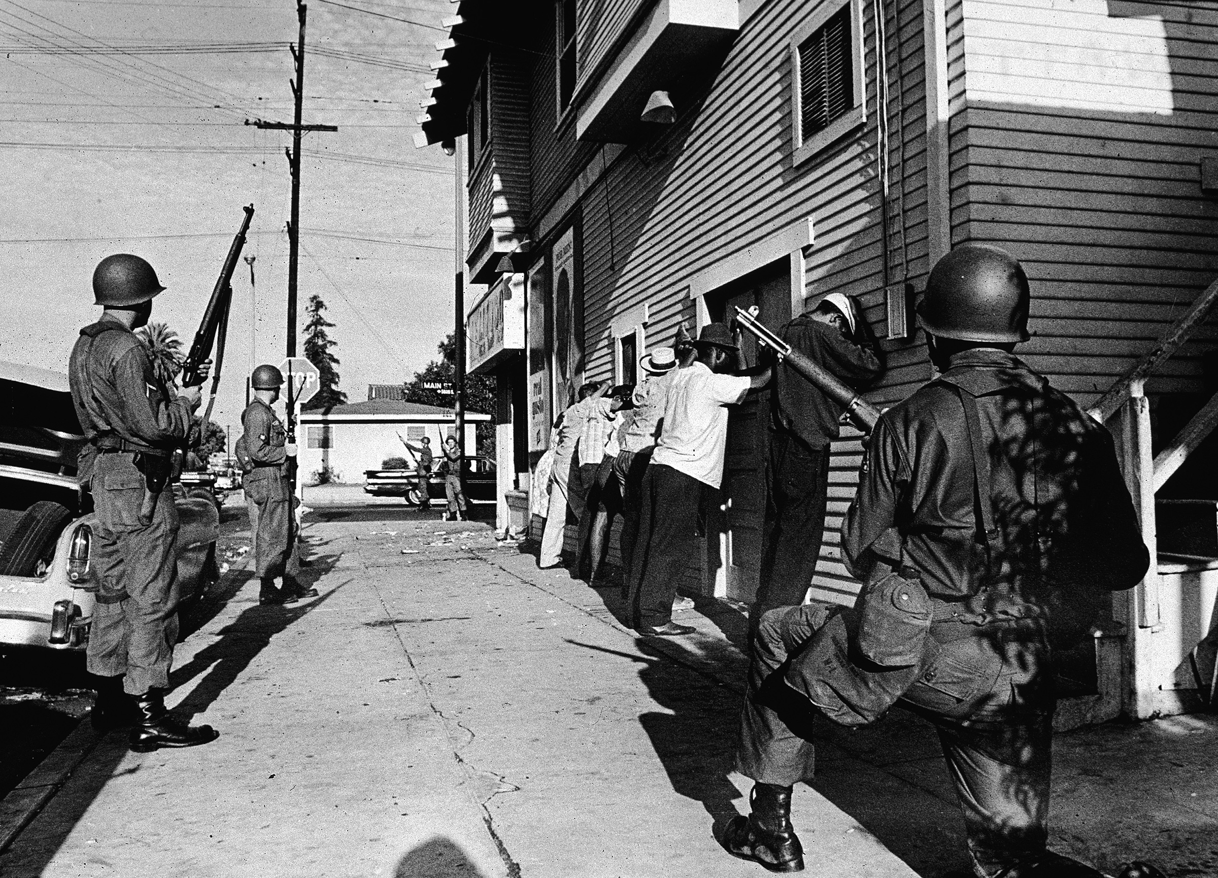 Armed National Guardsmen force a line of Black men to stand against the wall of a building during the Watts riots