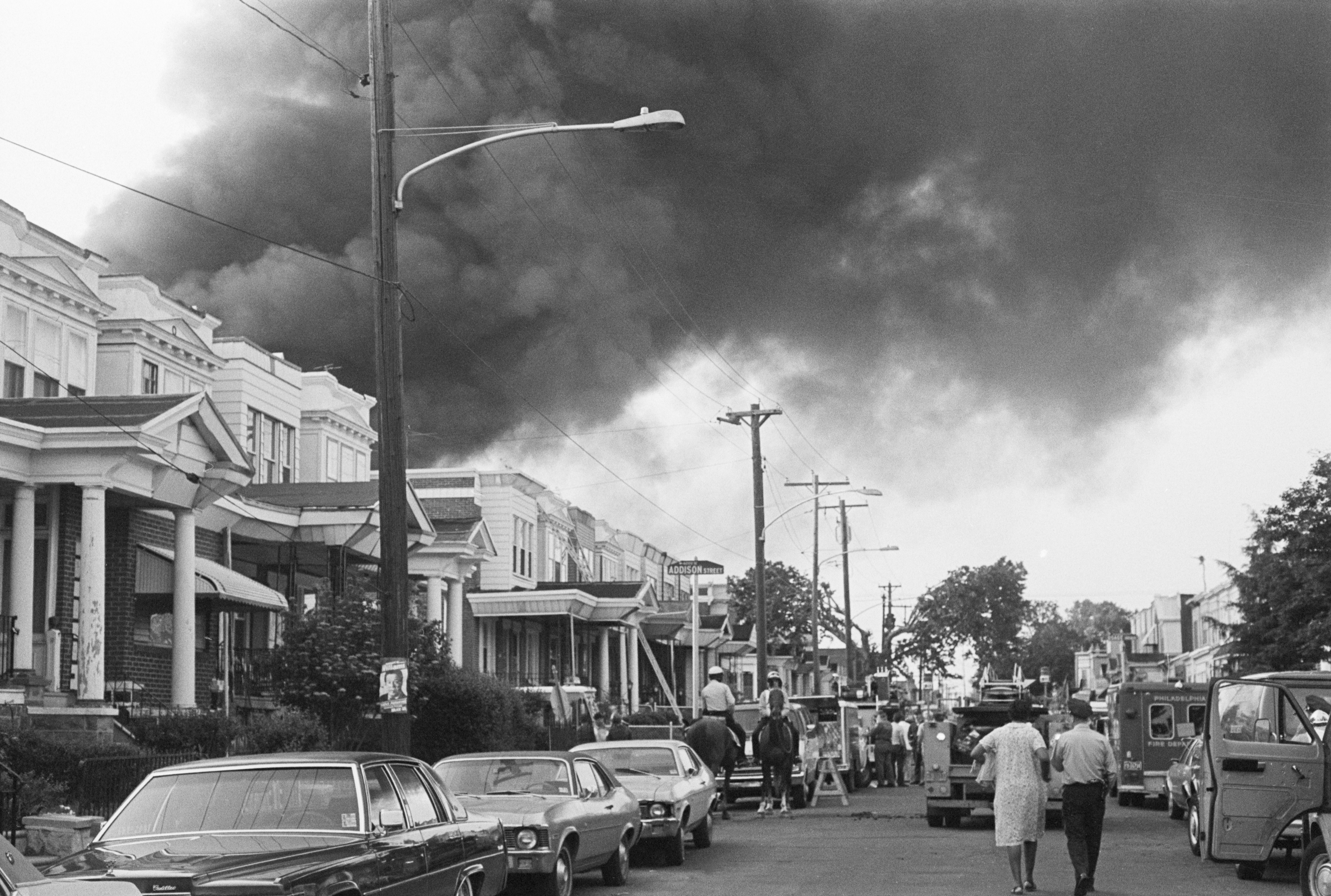 Smoke billows over rowhouses in West Philadelphia on May 12, 1985 after the police bombed a residence home to members of the Black liberation group MOVE. Police on horseback and emergency vehicles block off a street as residents walk towards the scene. (Bettmann Archive—Getty Images)