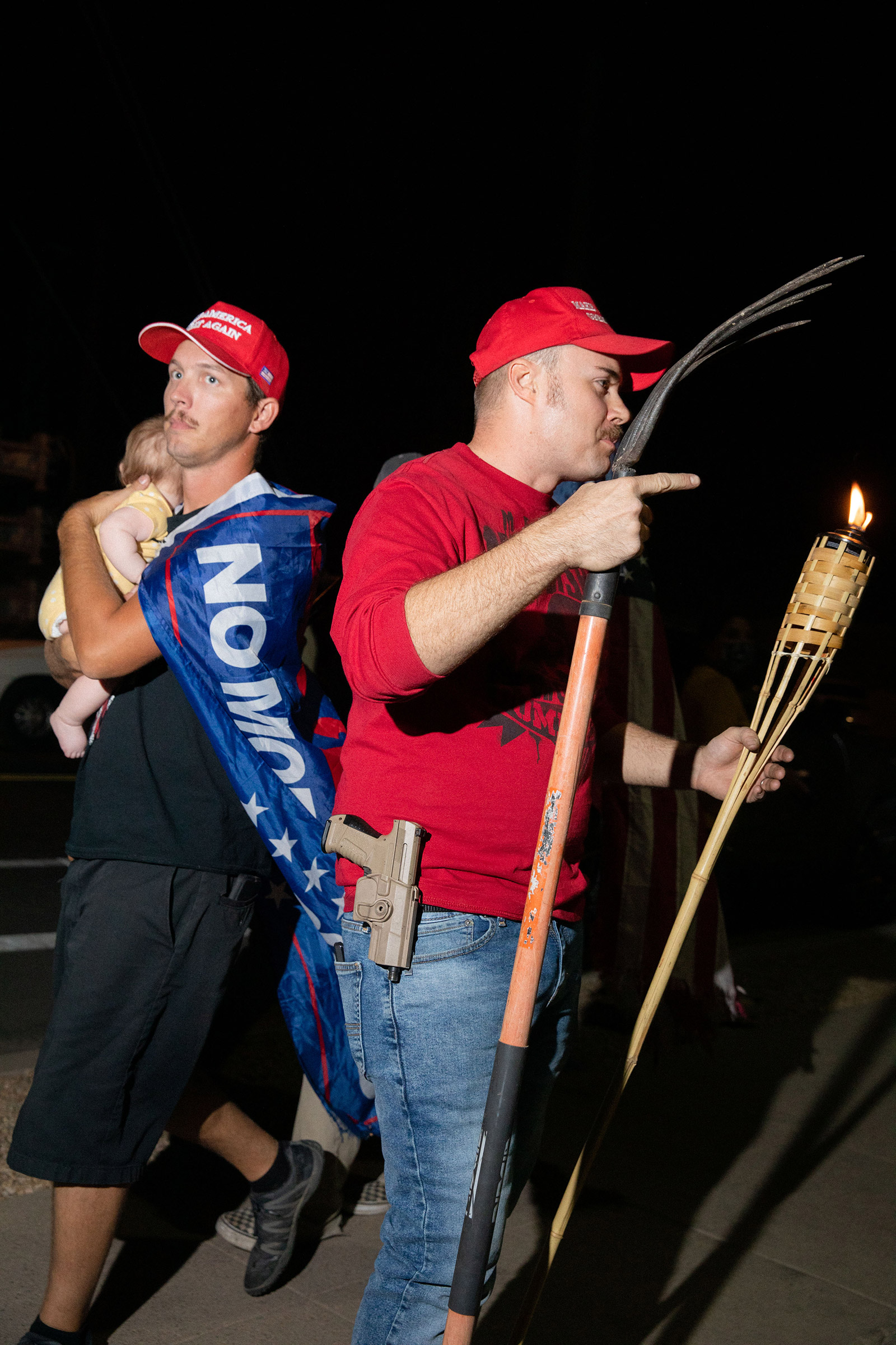 A father with child walks by a man with a rake, gun and tiki torch at the Maricopa County Elections office in Phoenix, on Nov. 5