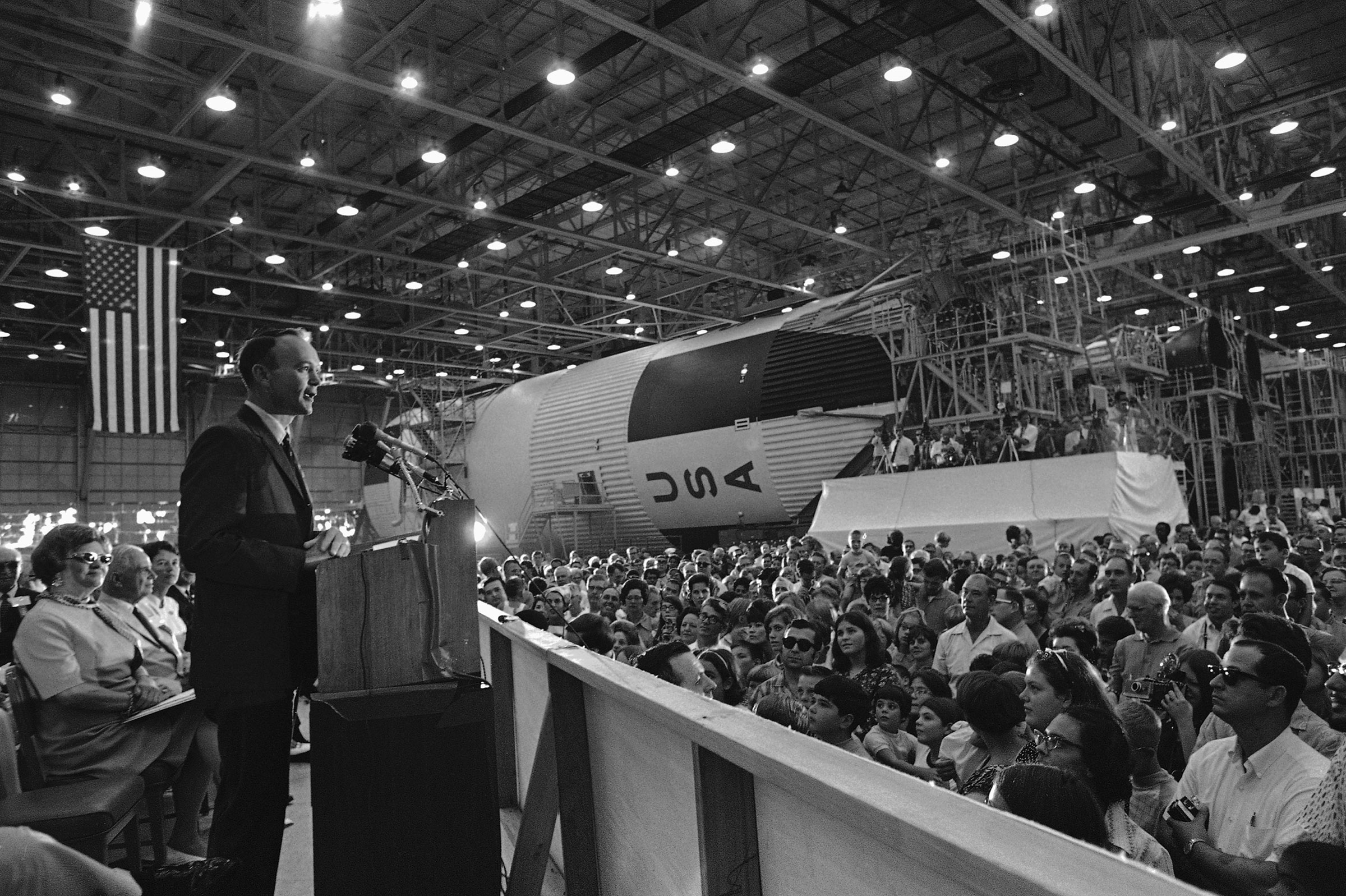 Astronaut Michael Collins addresses a crowd at the New Orleans Michoud Assembly Plant as a model of the giant Saturn 5 booster that sent the Apollo crew on their way to the moon rests in the background on Sept. 6, 1969.