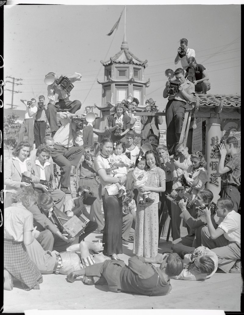 A mob of photographers descended upon China City, Los Angeles, when they heard that a prize contest is under way for the best amateur snaps of its people and its colorful nooks and corners. Pictured is a mob of candid camera fans shooting Mrs. Dorothy Siu (CQ) and Mrs. Chung Dat Loo as they hold little Gwendolyn Loo and Evelyn Loo on Oct. 1939