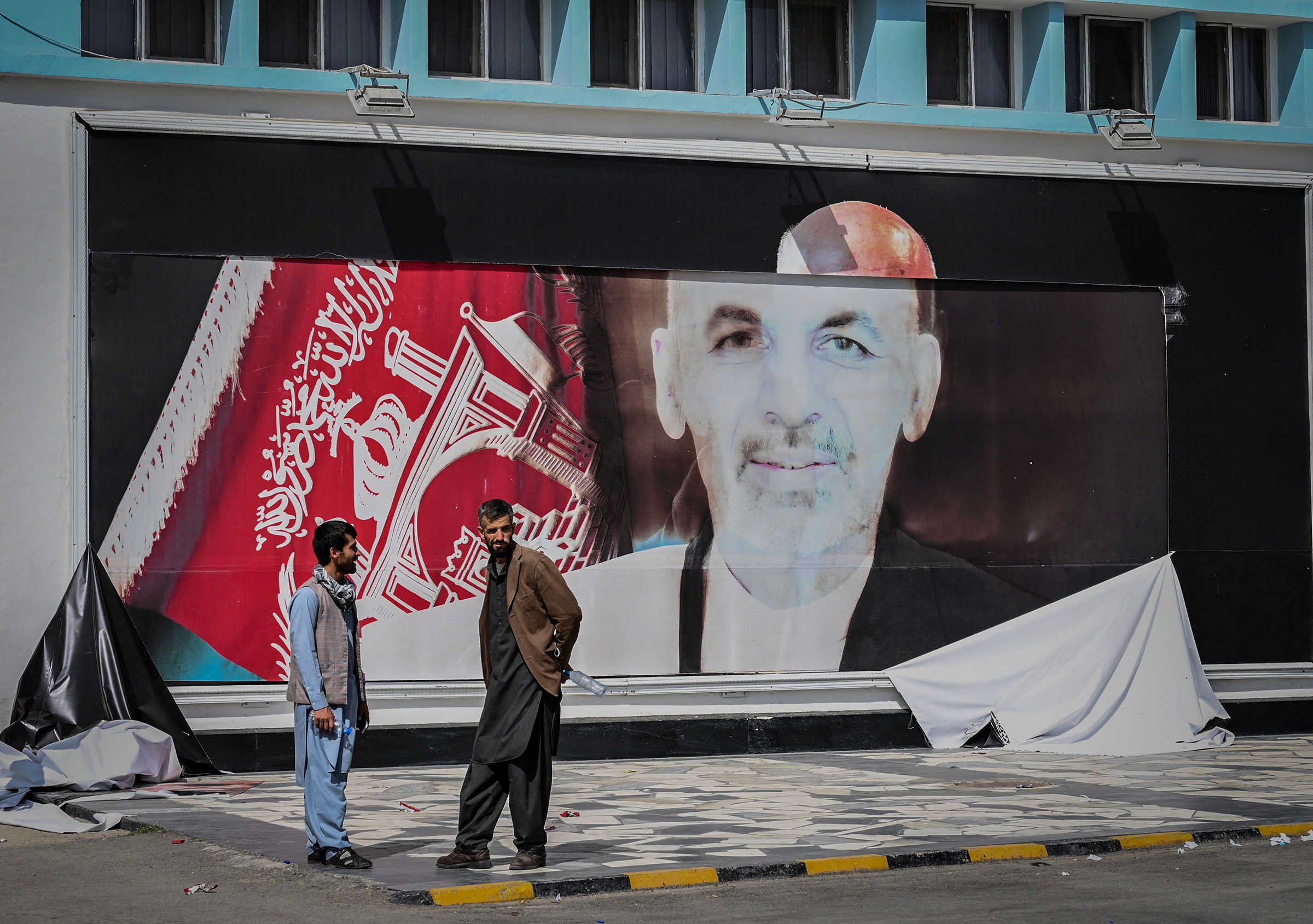 Men stand next to a torn poster of Afghan President Ashraf Ghani at the Kabul airport on Aug. 16, 2021, the day after Ghani fled the country and the Taliban gained control of the presidential palace.