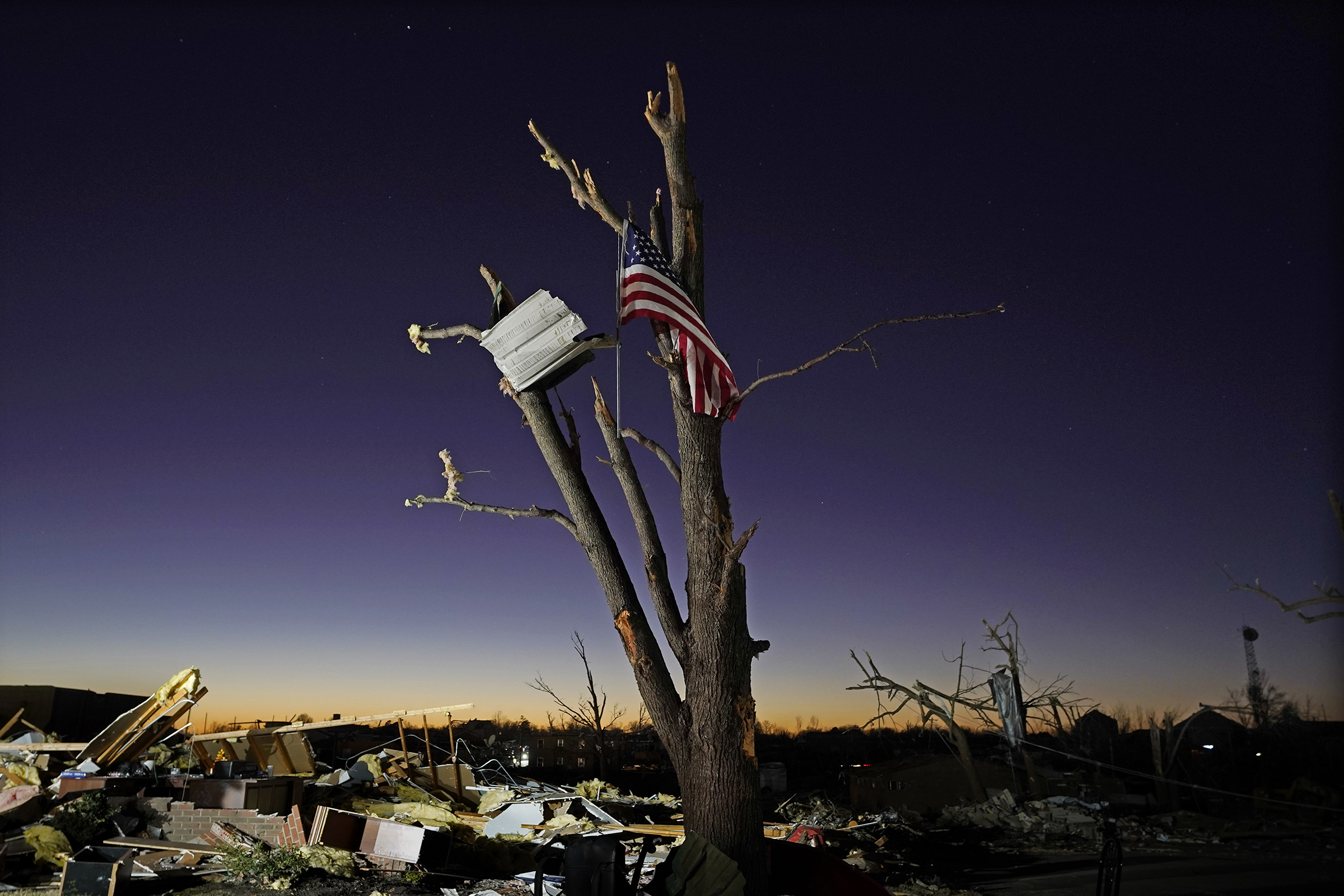 An American flag hangs from a damaged tree Sunday, Dec. 12, 2021, in Mayfield, Ky. Tornadoes and severe weather caused catastrophic damage across several states Friday, killing multiple people. (AP Photo-Mark Humphrey)