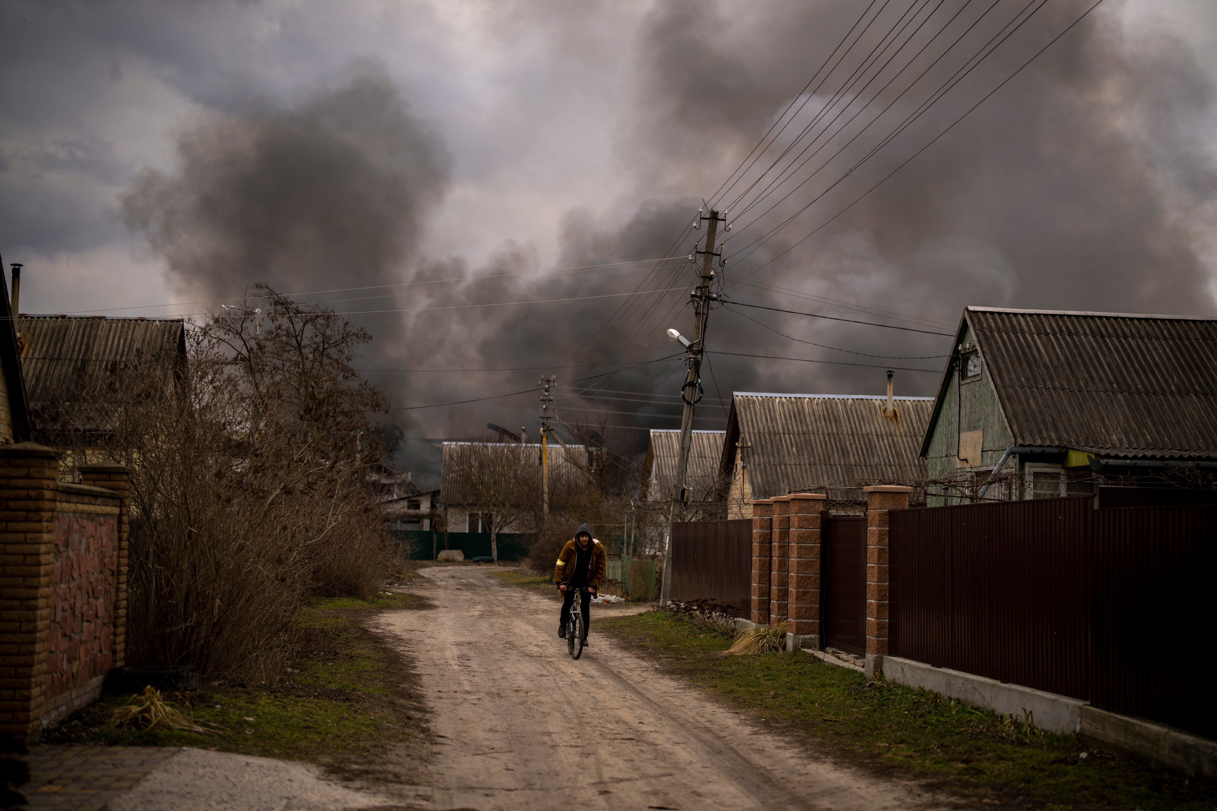 A Ukrainian man rides his bicycle near to a factory and a store burning after been bombarded in Irpin, on the outskirts of Kyiv, Ukraine, Sunday, March 6, 2022. (AP Photo/Oleksandr Ratushniak)