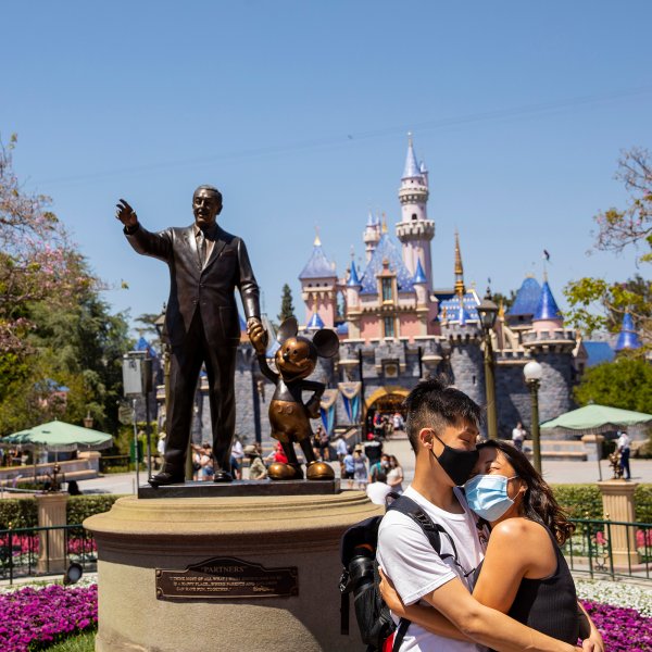 A masked couple poses for photos in front of a statue of Walt Disney and Mickey Mouse, with Sleeping Beauty Castle behind, at Disneyland Resort in Anaheim, CA, as visitors return to the park with covid-safety restrictions in place, including the park only being at 25% capacity, Monday, May 3, 2021.