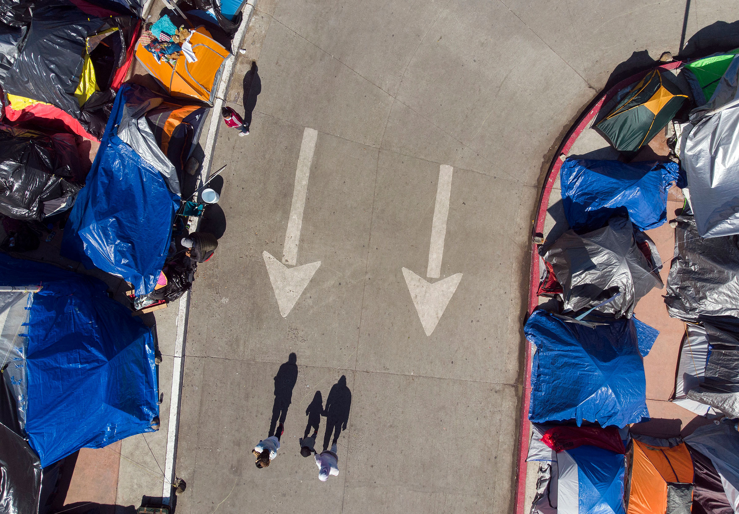 Aerial view of three migrants casting their shadow on the pavement by a migrants camp where asylum seekers wait for US authorities to allow them to start their migration process outside El Chaparral crossing port in Tijuana, on March 17, 2021.