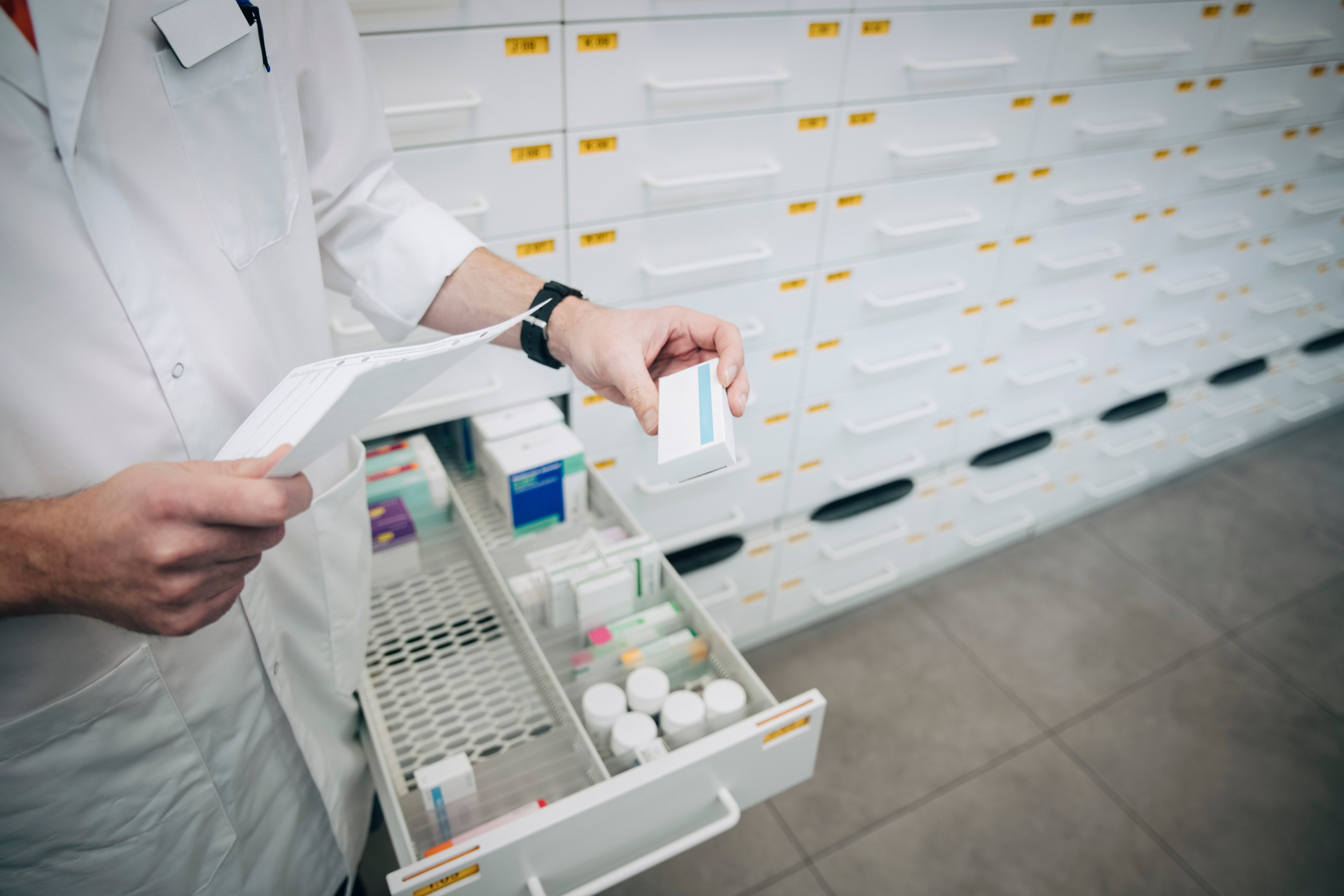 Midsection of mature male pharmacist holding prescription paper and medicine standing by drawer at store