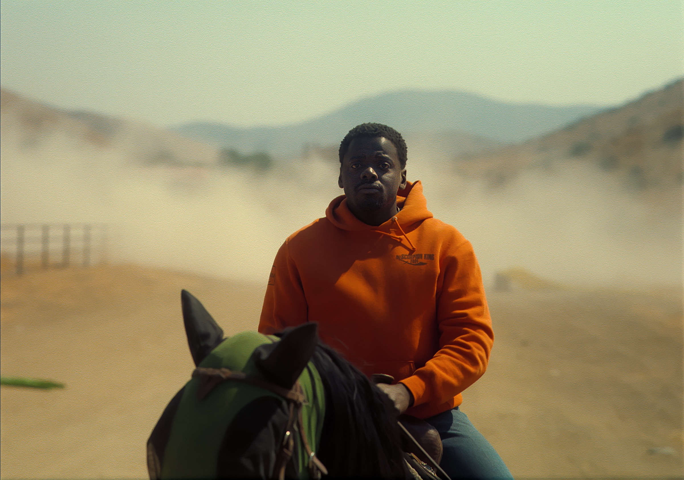 Jordan Peele in an orange hoodie, on horseback, rides toward the camera