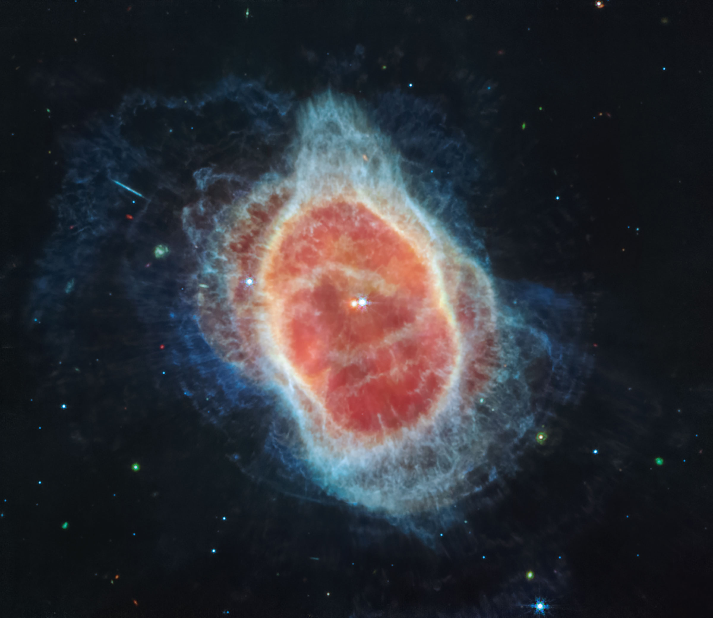 A planetary nebula seen, by Webb’s MIRI, against the blackness of space, with points of starlight behind it. Plumes of glowing bright blue gas radiate out from the oval-shaped nebula. Two separate ovals of reddish pink gas appear stacked on top of each other, inside the lacy blue gas clouds. In the center of the nebula, two stars glow close to each other. One star looks more red, while the other appears more yellow. (NASA, ESA, CSA, STScI)