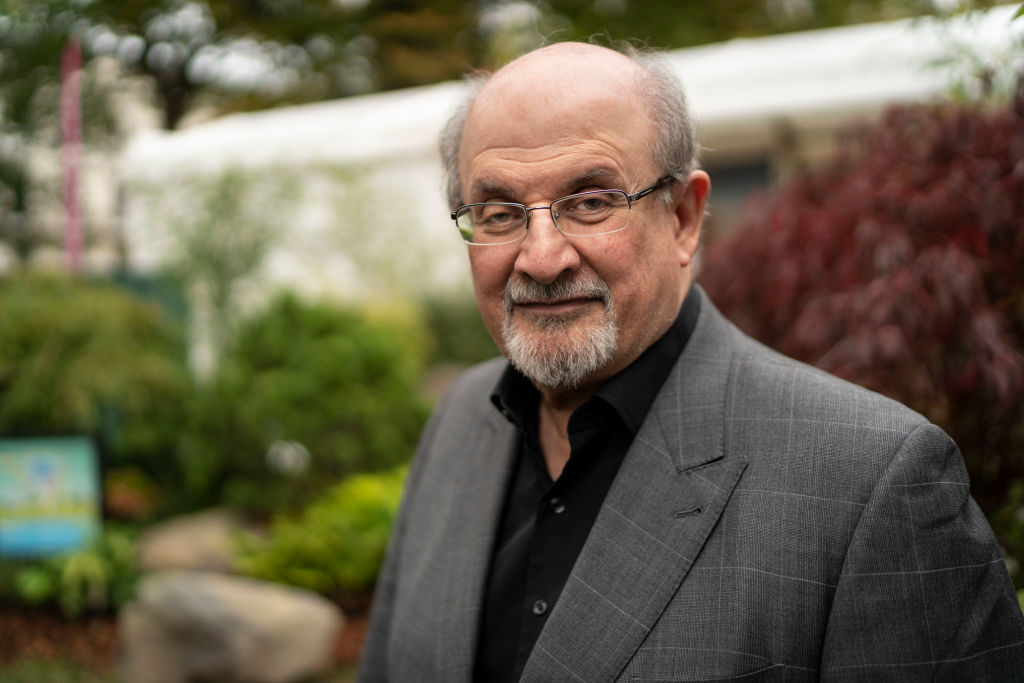Salman Rushdie smiles gently at the camera in a gray suit