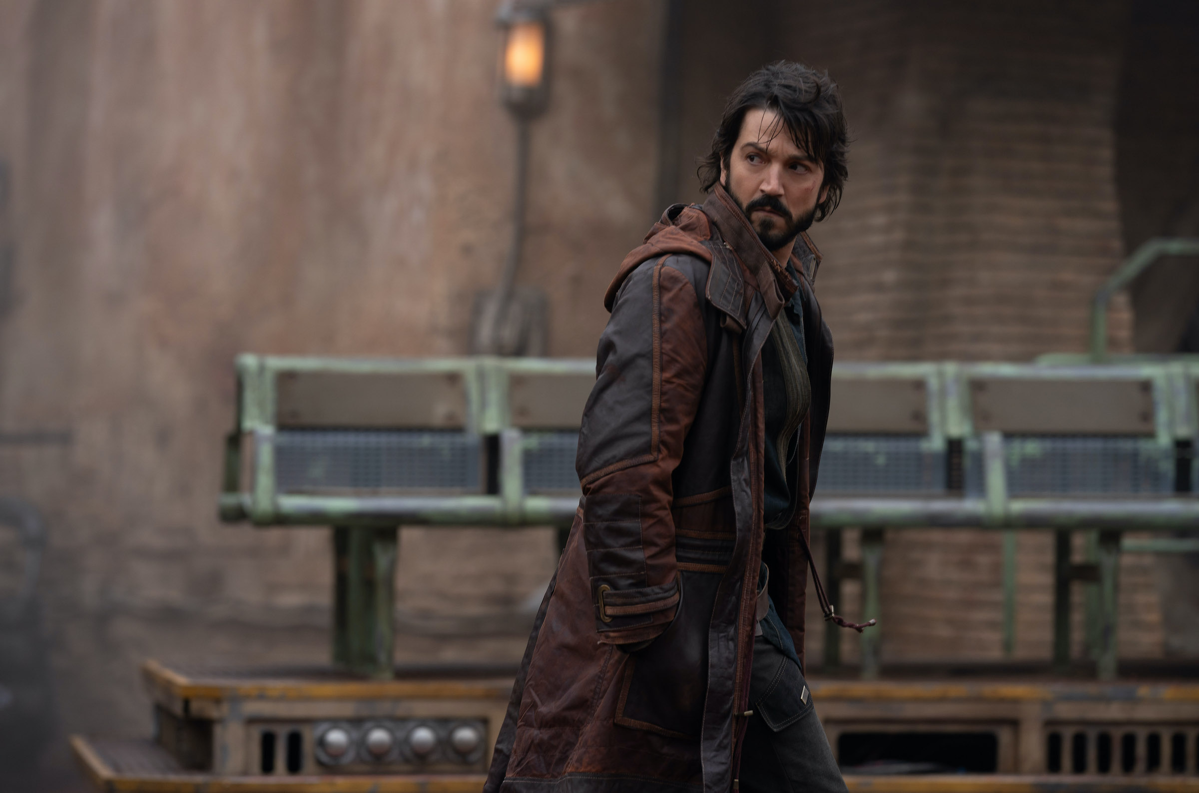 Diego Luna, dressed in a long brown coat, walks toward the right of the frame, looking behind him.